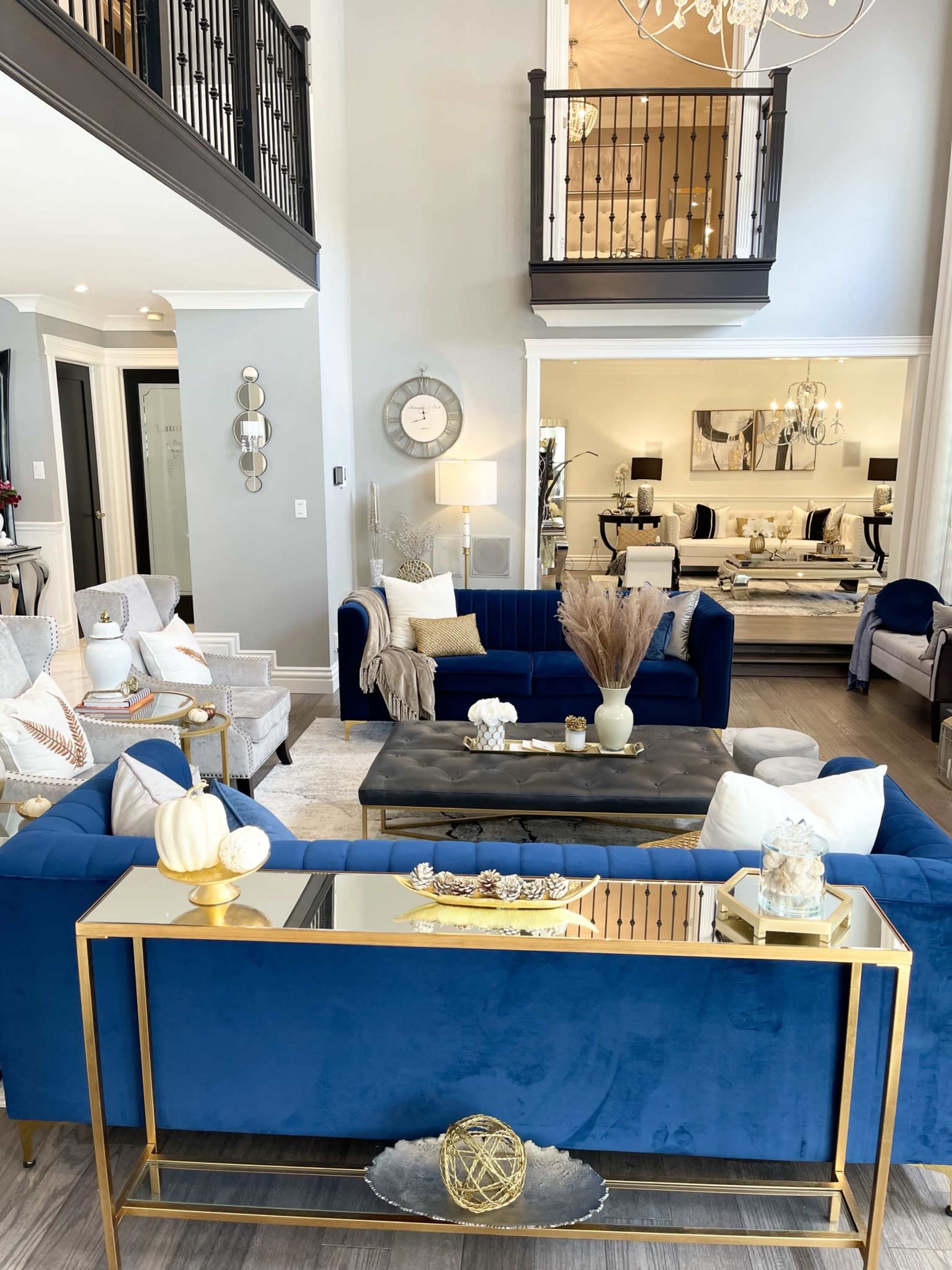 A modern living room with blue velvet sofas, a glass-top coffee table, and decorative elements like vases and a mirror, set in a spacious area with high ceilings and a balcony above.