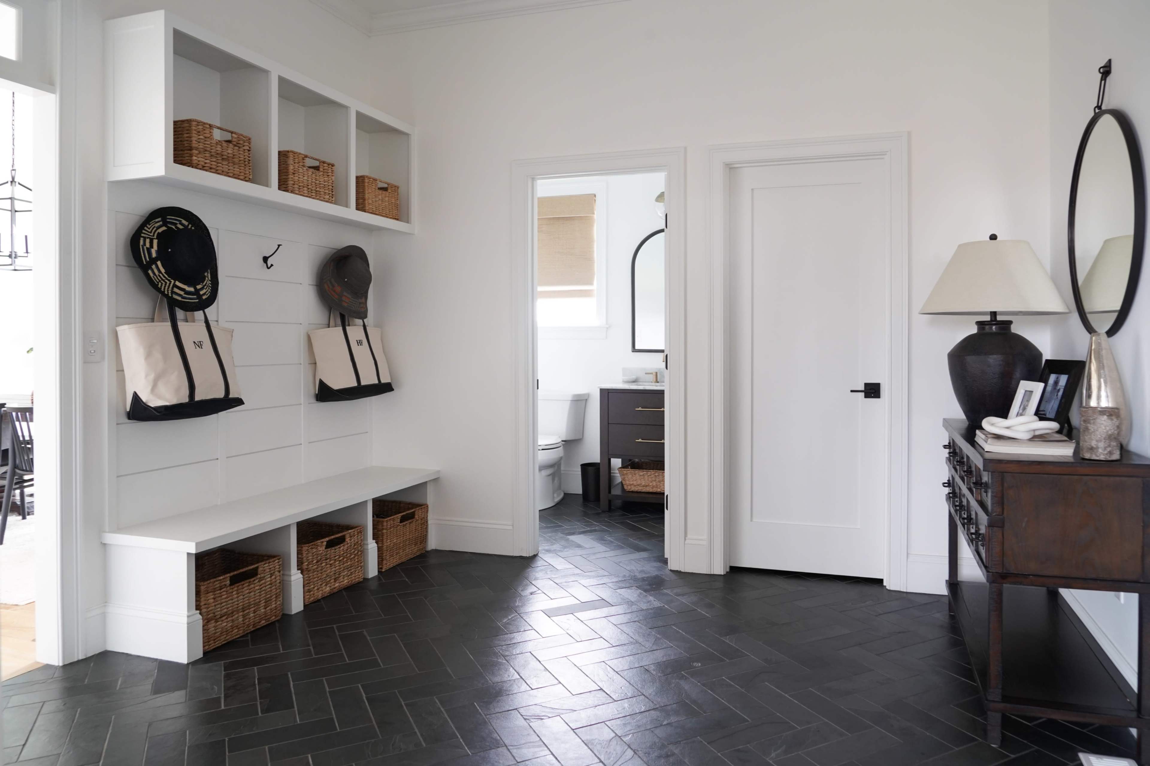 A modern entryway with a bench, wall-mounted hooks, and storage baskets, leading to a bathroom and featuring a mirror and a lamp on a console table.