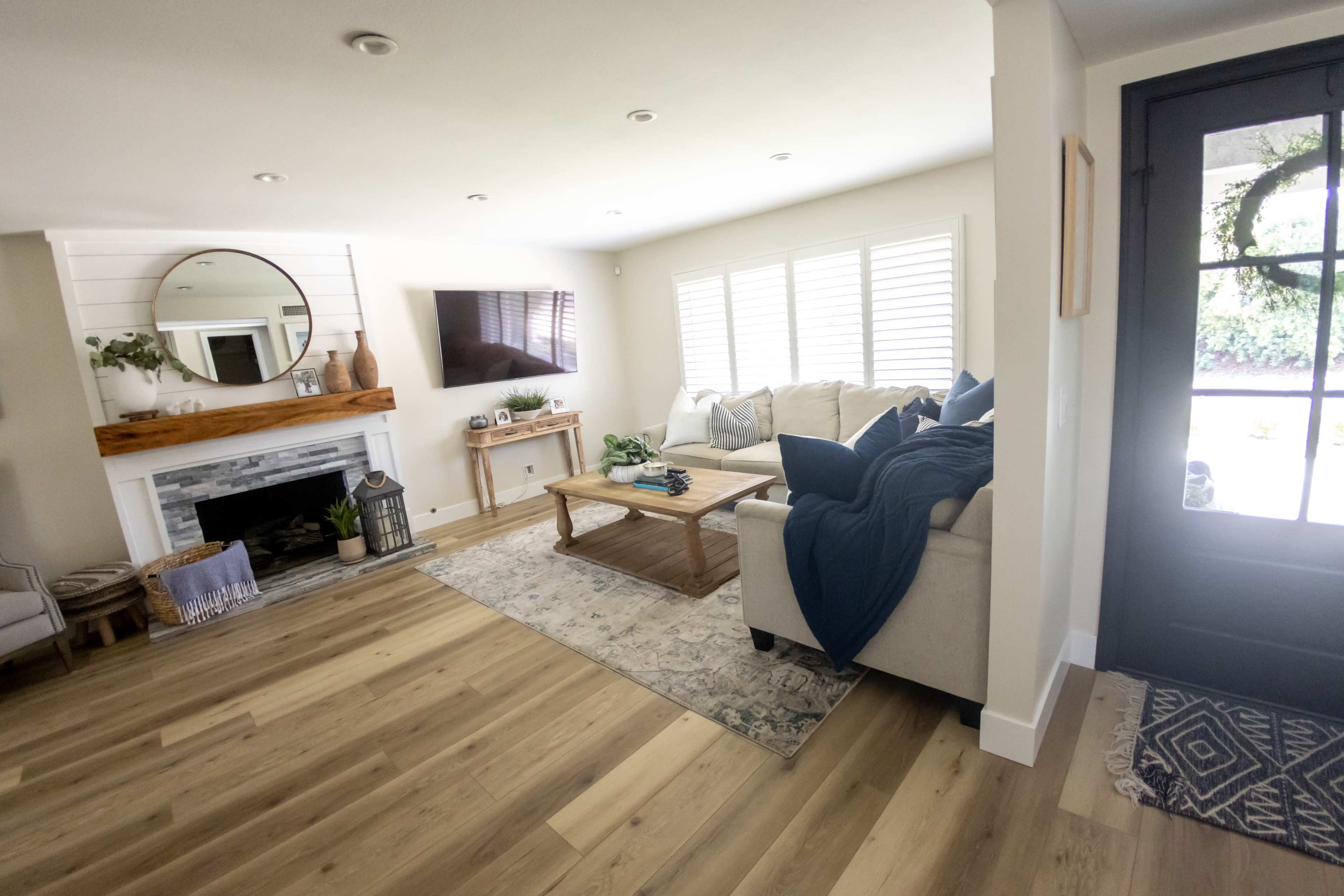 The image shows a cozy living room with a large sofa, a wooden coffee table, a television mounted on the wall, and a front door to the right.