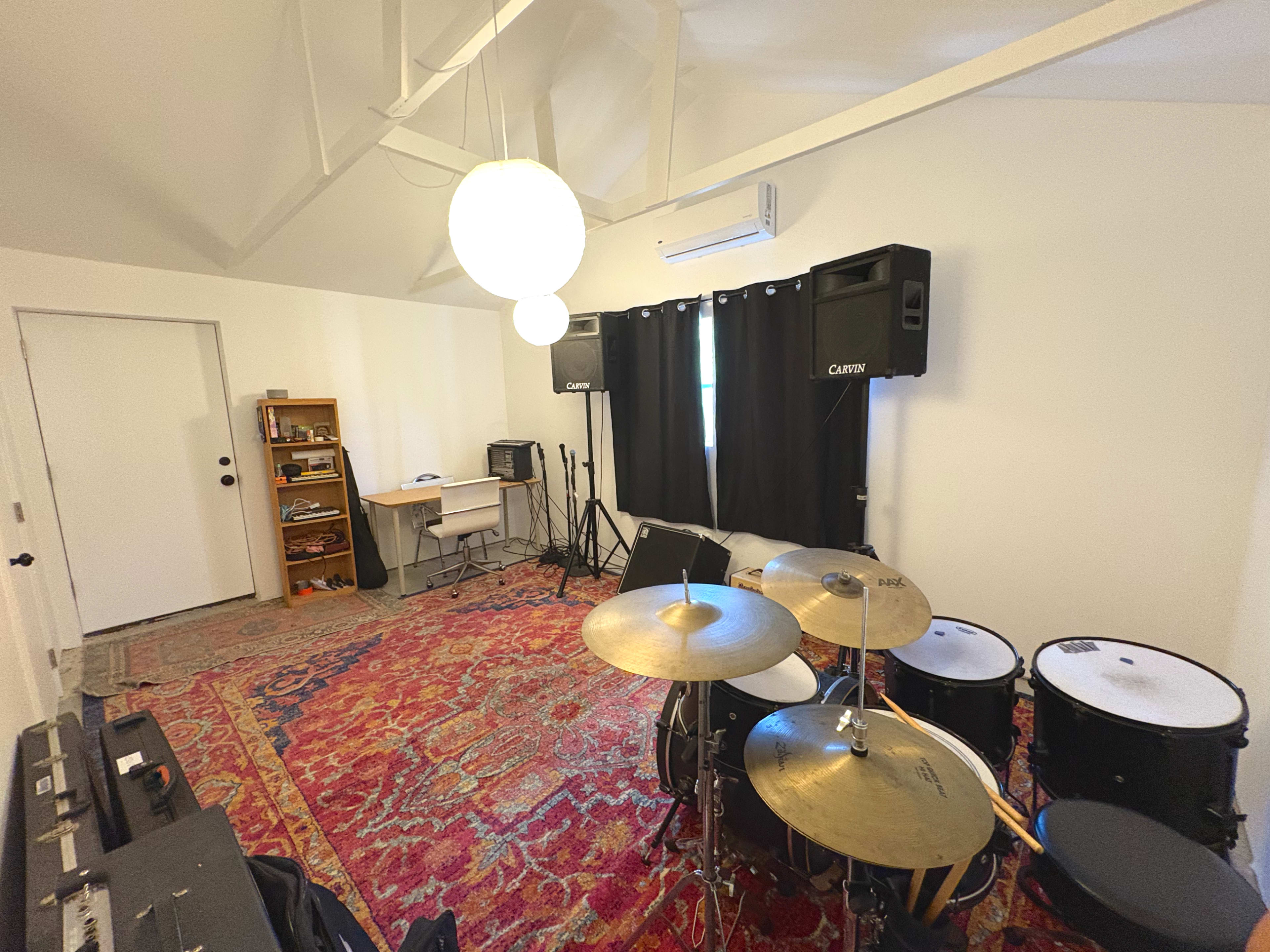 The image shows a music rehearsal room with drum equipment, amplifiers, a desk, and a decorative rug.