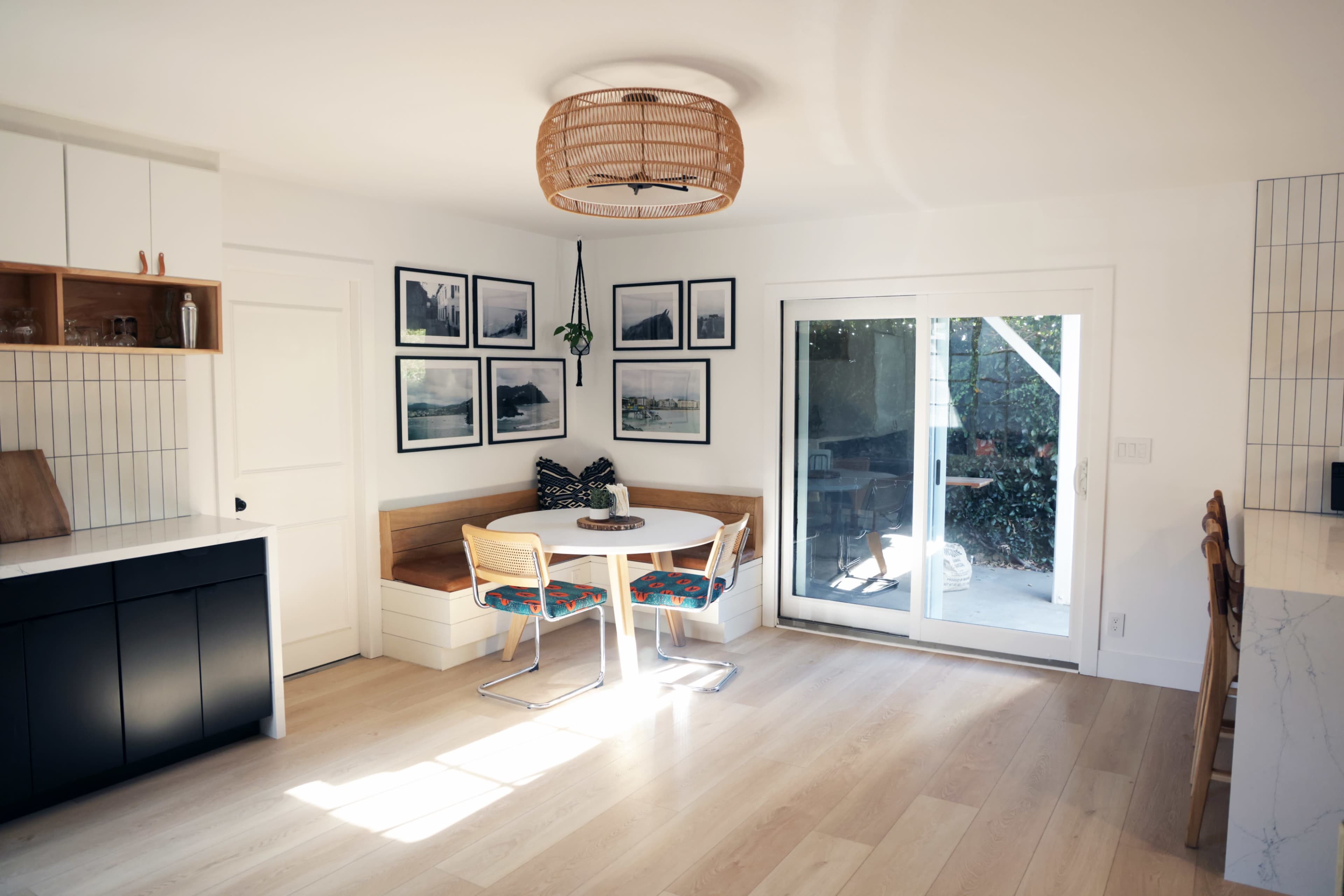 A bright kitchen features a circular dining table with colorful chairs and a built-in bench, surrounded by framed photographs and a sliding glass door leading to an outdoor area.