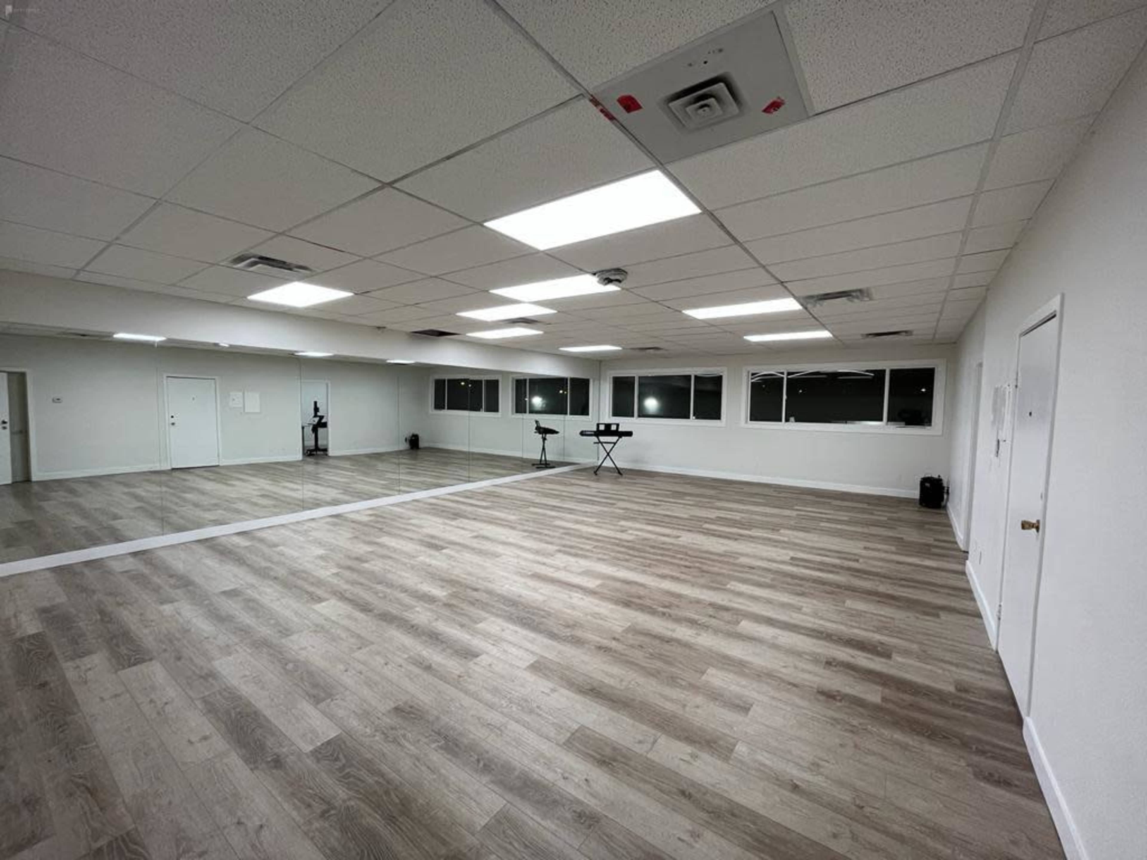 An empty room with wooden flooring, large windows, and mirrors on one wall, designed for activities like dance or fitness.