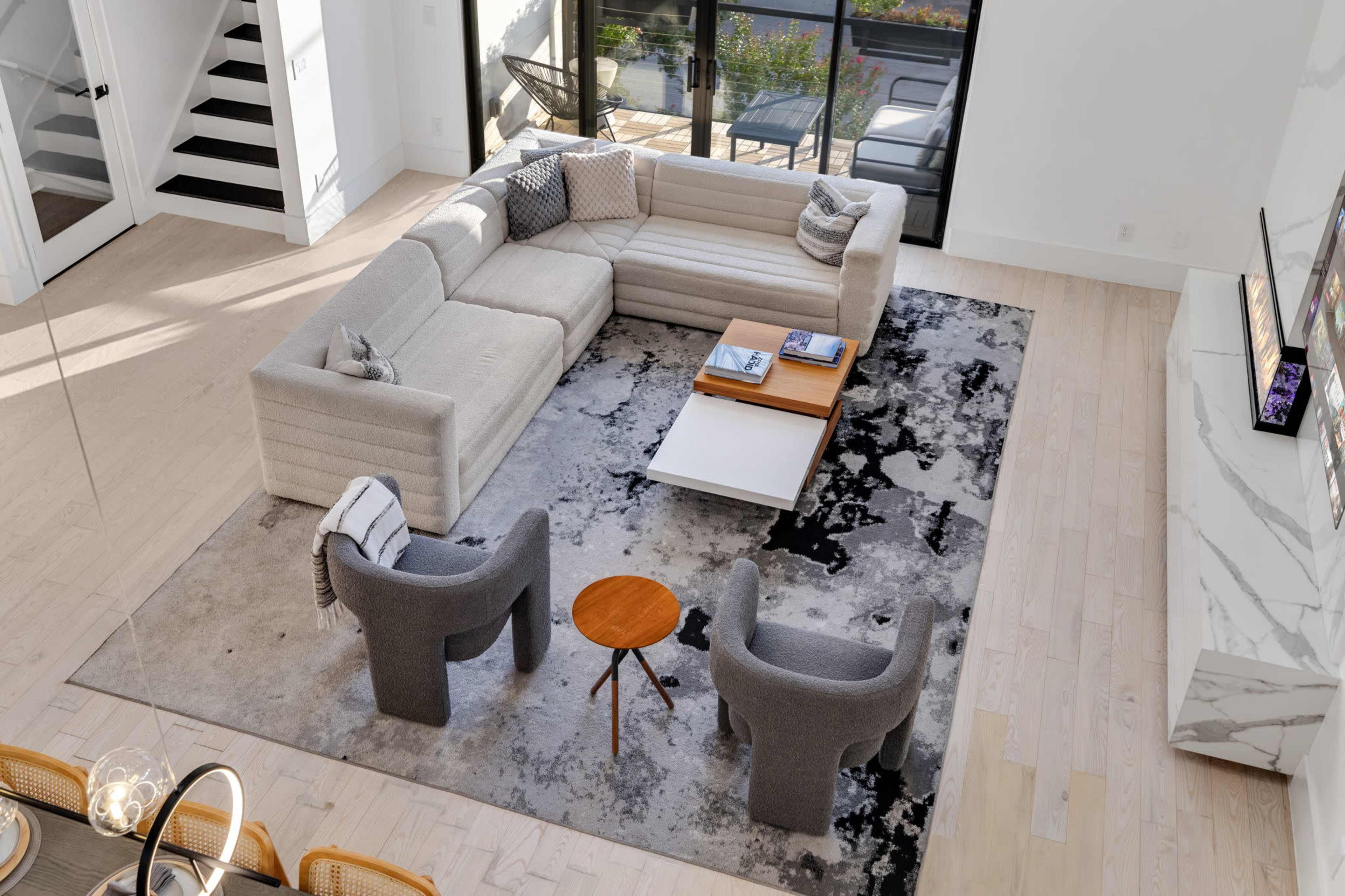 The image showcases a modern living room with a light-colored sectional sofa, two gray accent chairs, a wooden side table, and a rug with abstract patterns, all arranged around a coffee table.