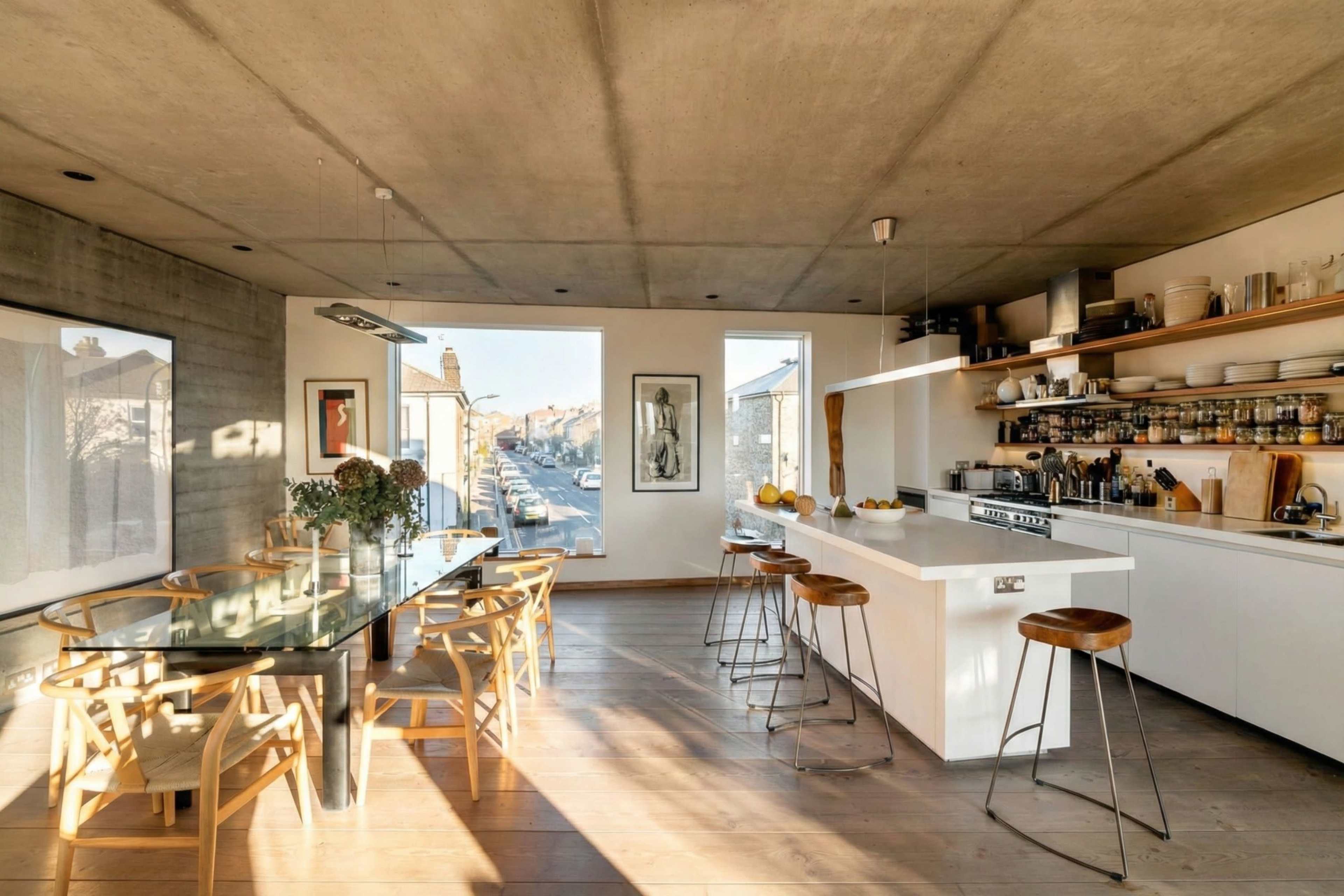 The image shows a modern kitchen and dining area with a concrete ceiling, large windows, a glass dining table, wooden floors, and a minimalist kitchen counter with bar stools.