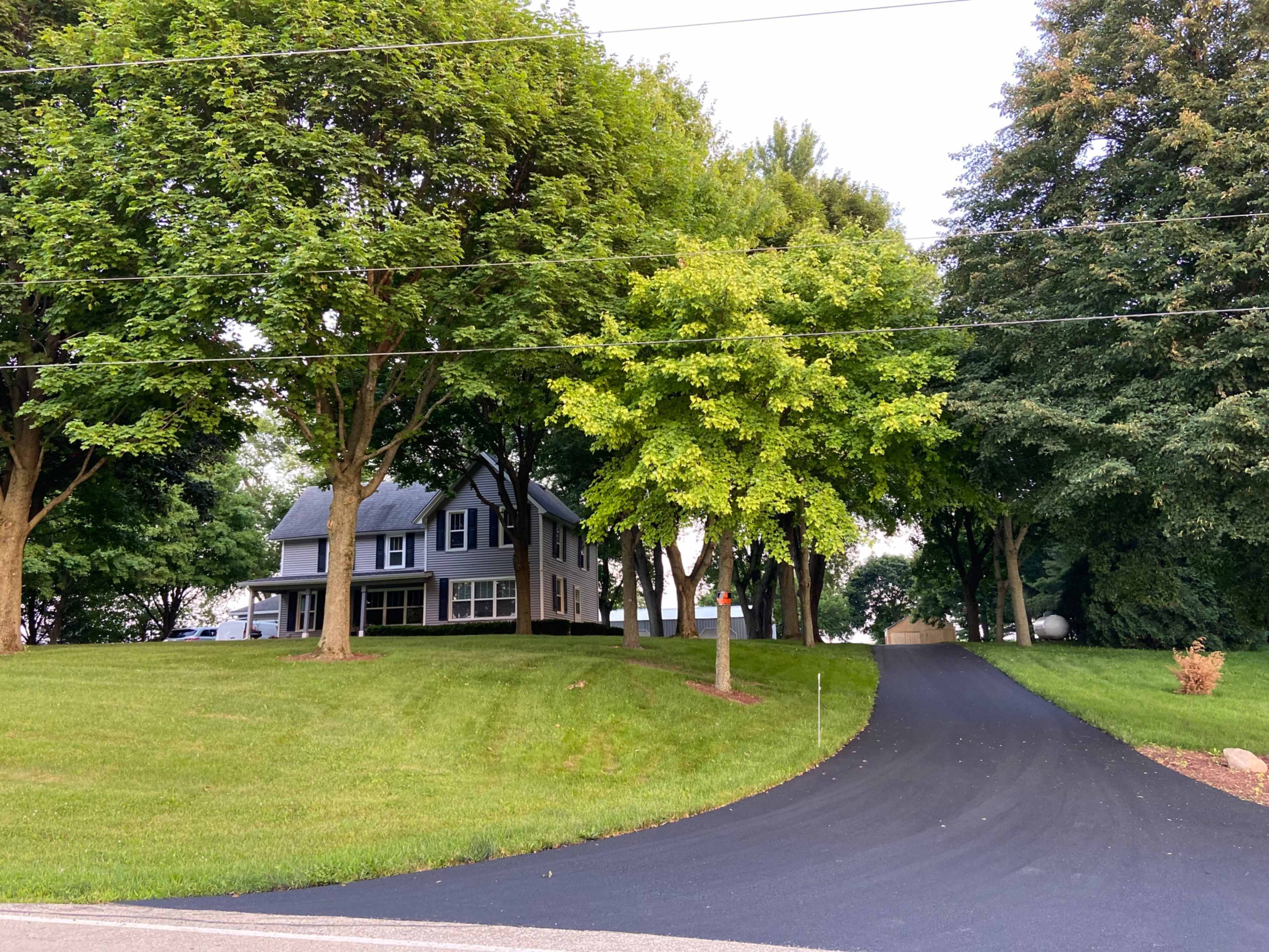 A grey house is situated on a grassy lot, surrounded by several trees, with a newly paved driveway leading to it.