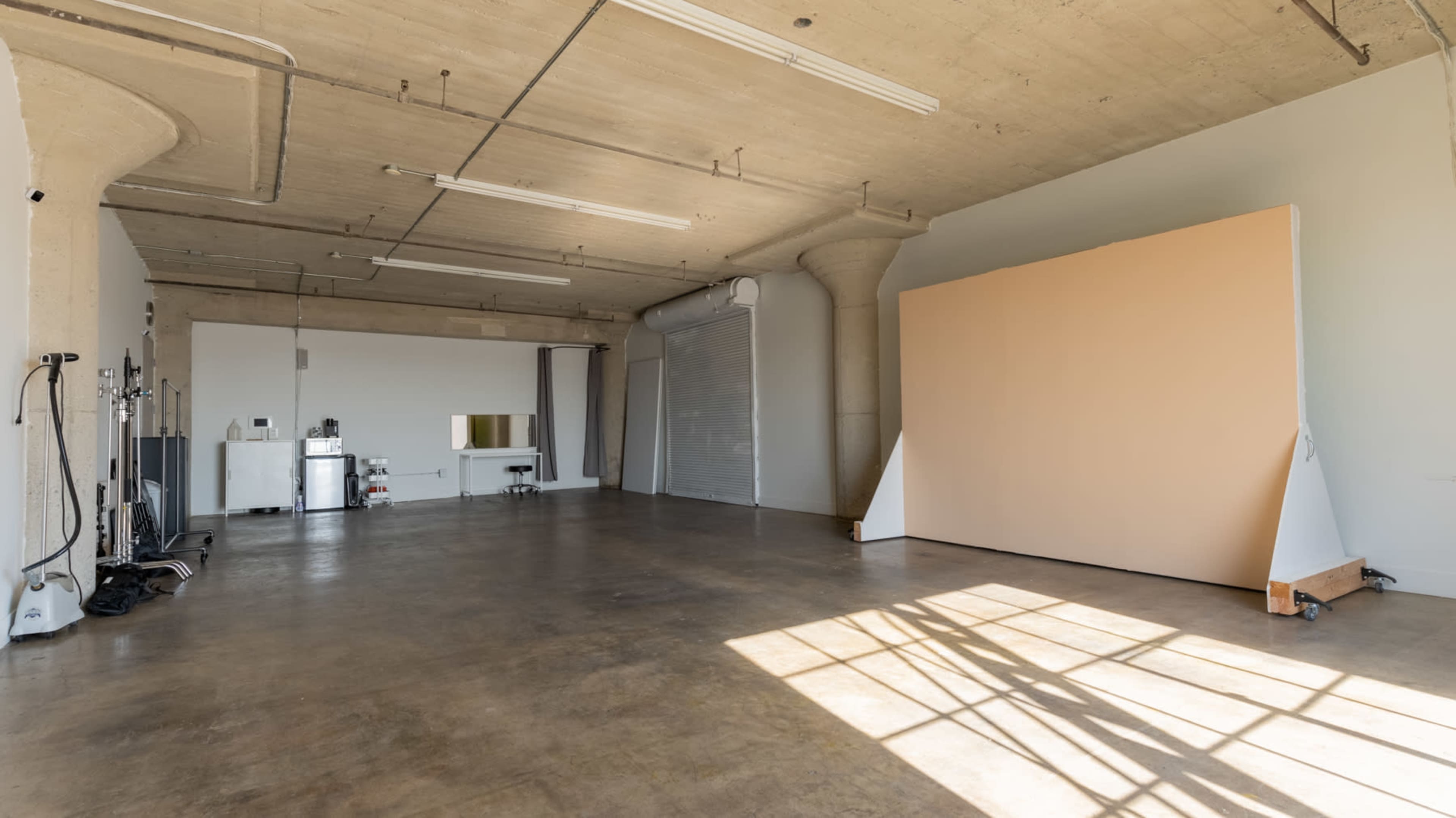 A spacious, empty studio with concrete floors, a large blank backdrop, and a kitchenette area in the background.