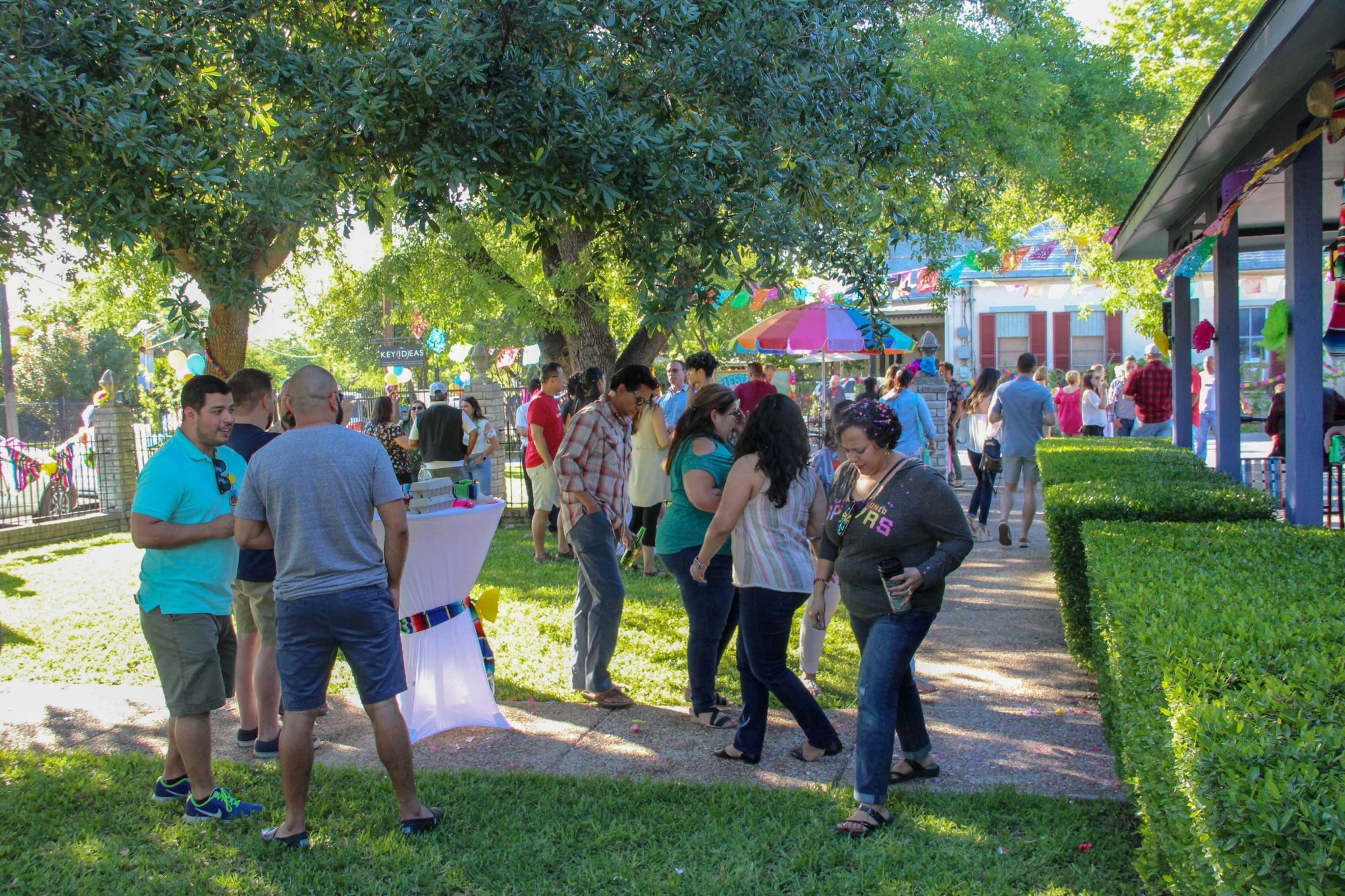 A group of people is gathered outdoors, mingling along a pathway lined with greenery during a festive event.