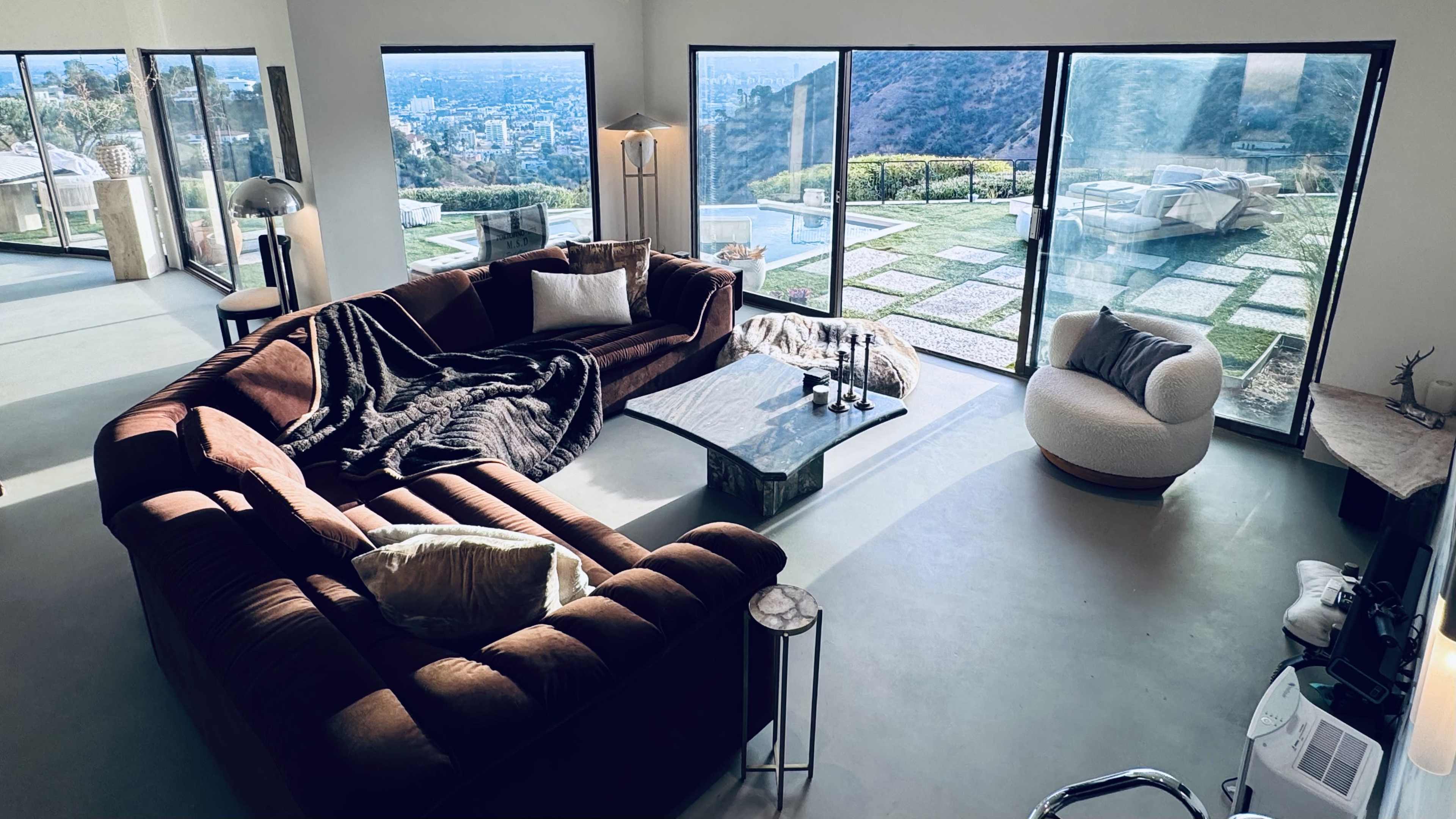A spacious living room features a large brown sectional sofa, a round chair, and expansive glass doors that offer a view of a hillside landscape.