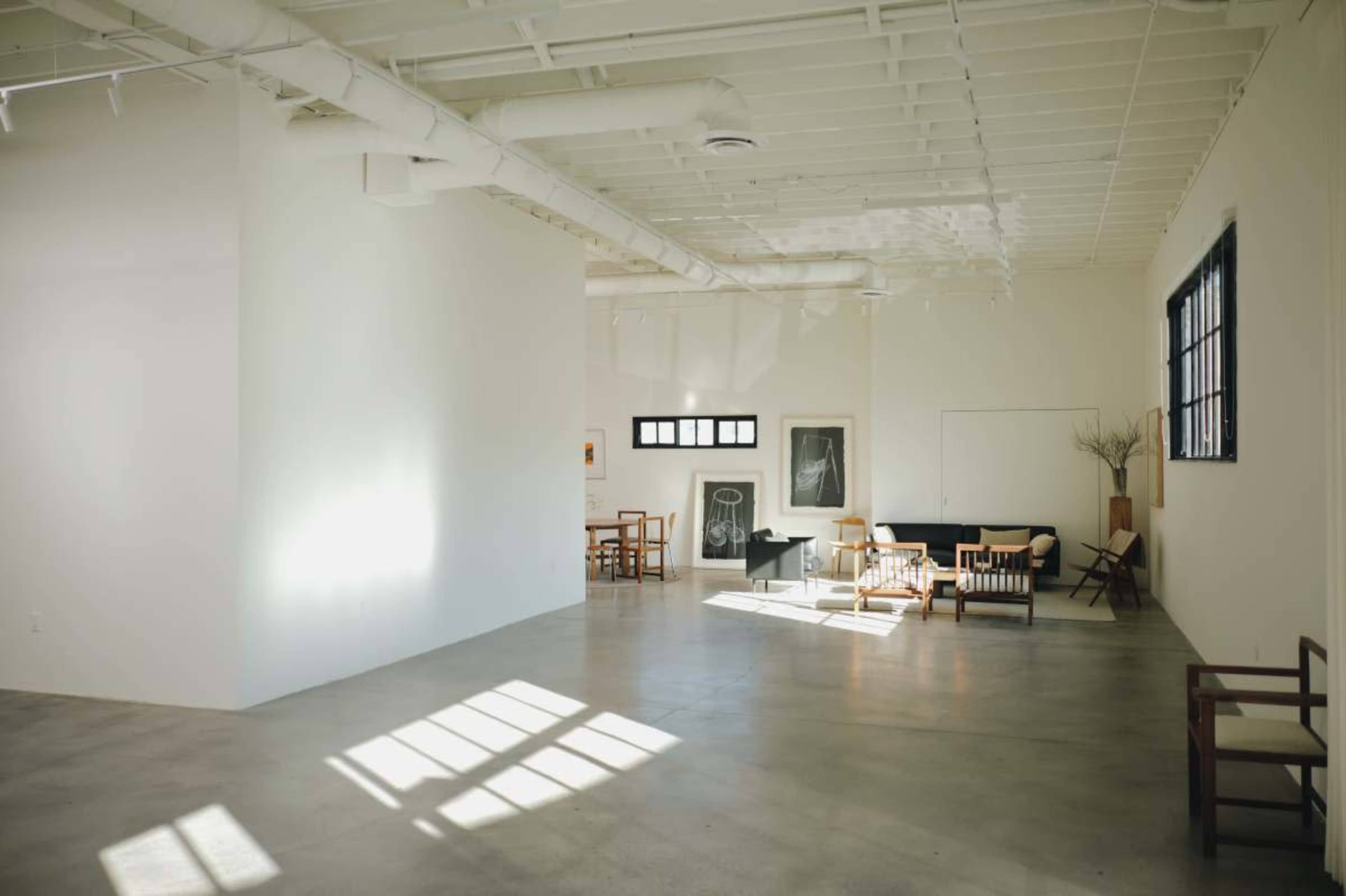 The image shows a spacious, minimalist room with concrete flooring, large windows, and a few pieces of furniture arranged in the space.