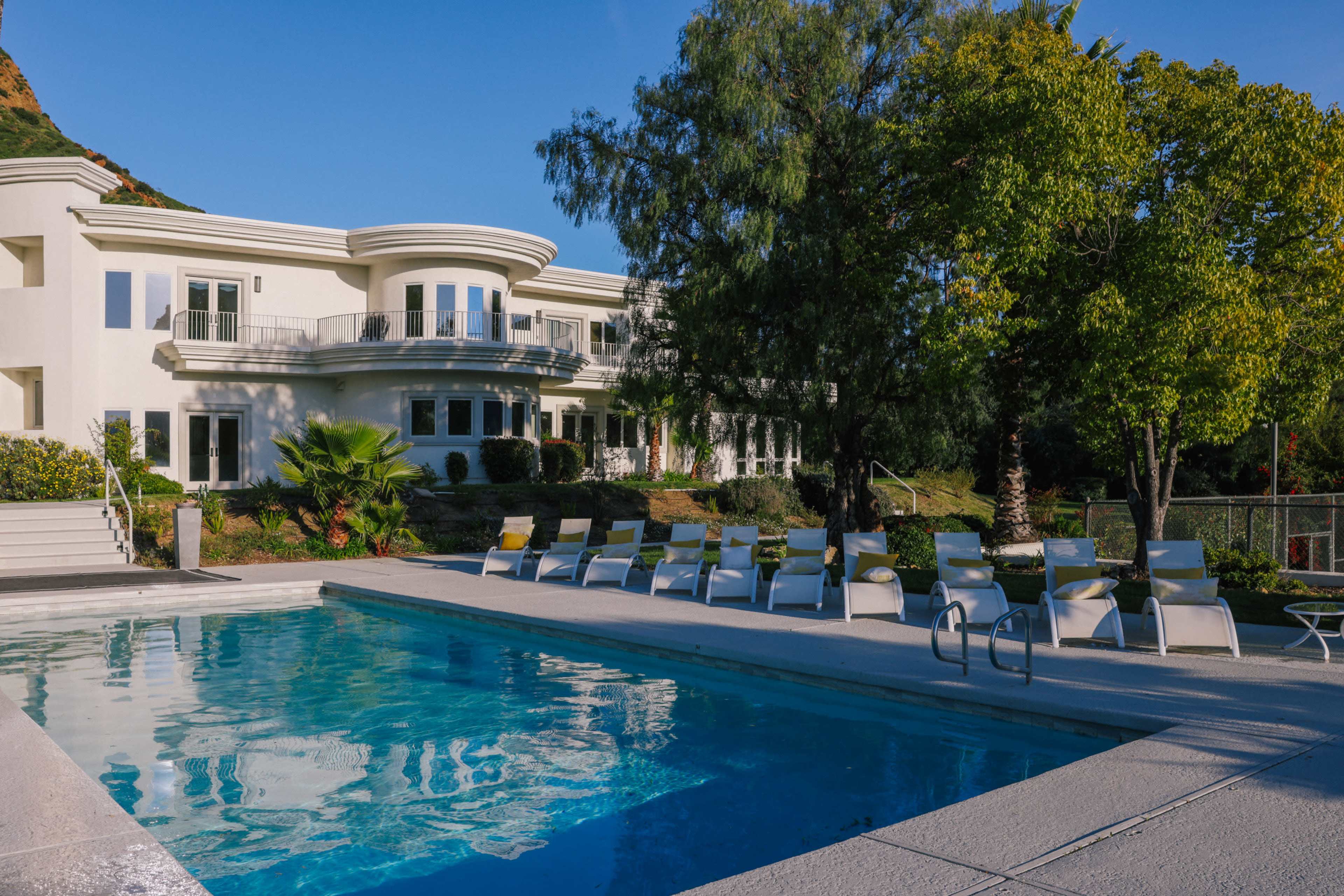 A modern, two-story house with a curved design overlooks a swimming pool surrounded by lounge chairs and palm trees.