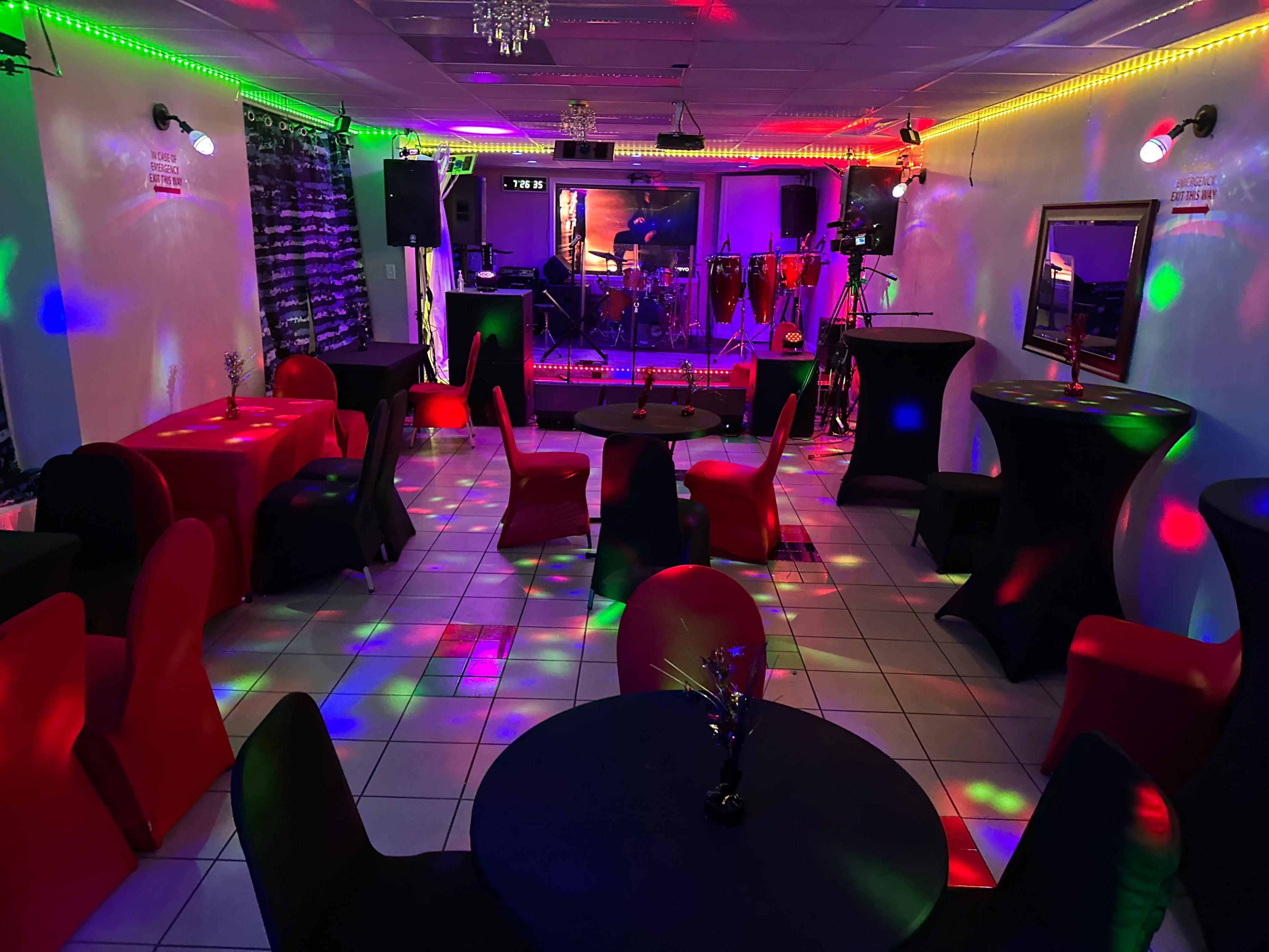 A brightly lit event space with colorful lights, tables covered in black and red cloths, and a stage set up for live music in the background.