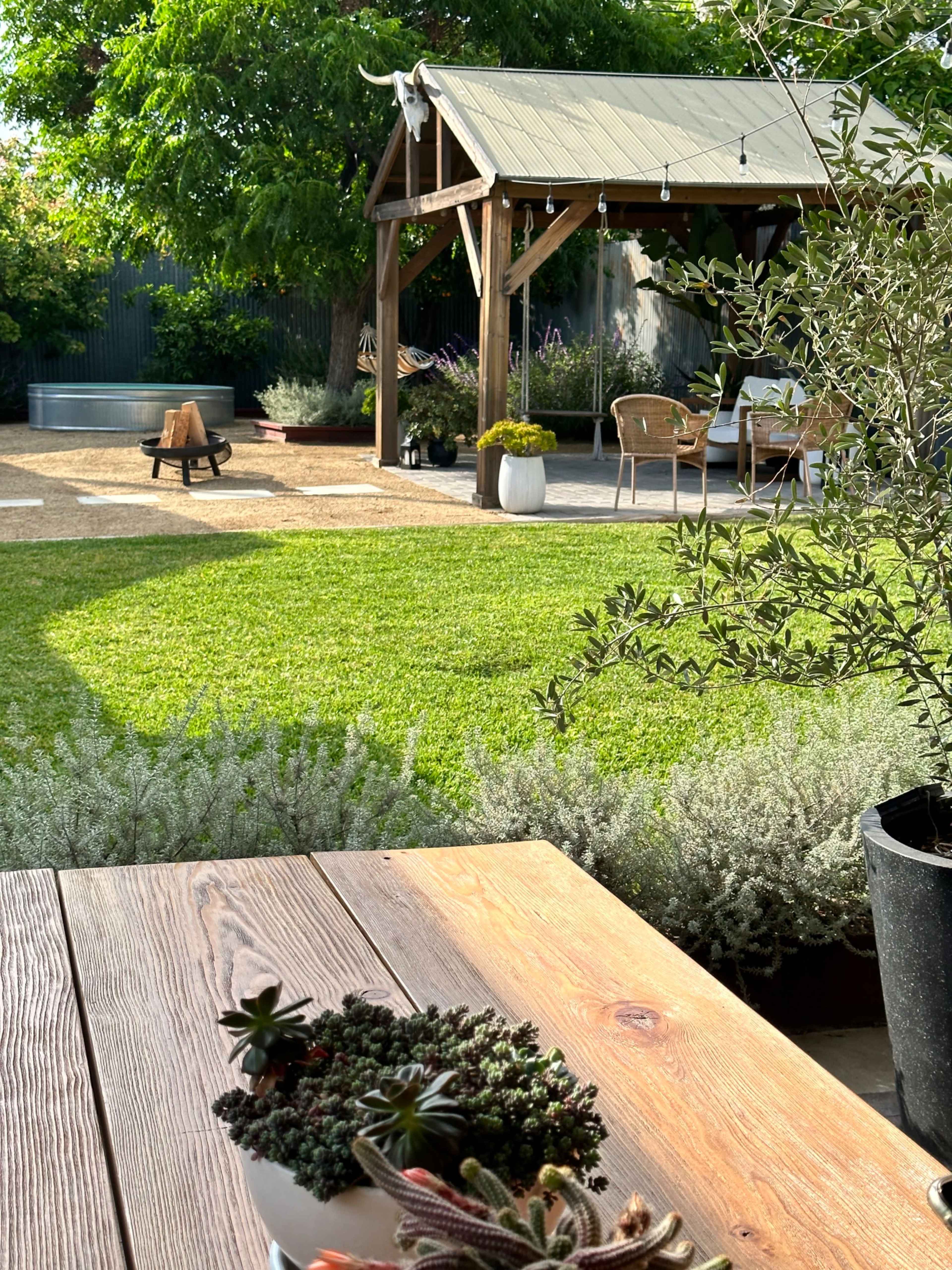 A well-maintained garden featuring a wooden gazebo, seating area, and a large grassy lawn with potted plants.