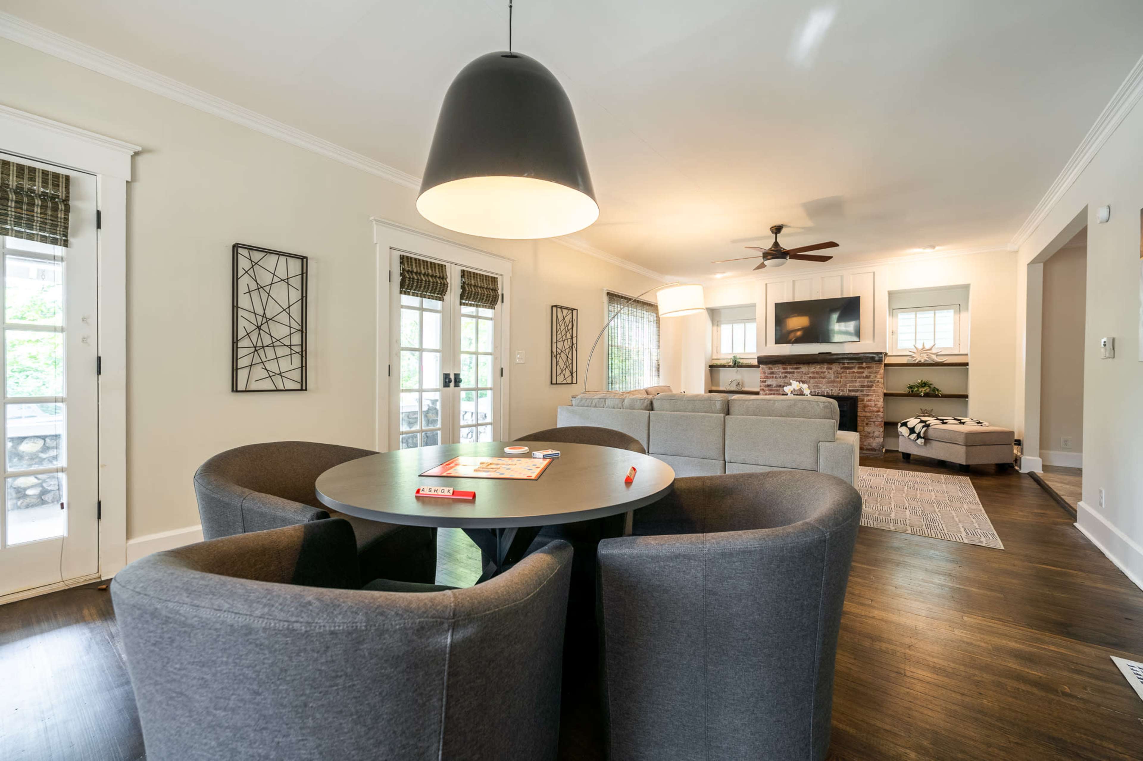 A modern living room features a circular table with four gray chairs, surrounded by natural light from large windows and a spacious couch in the background.