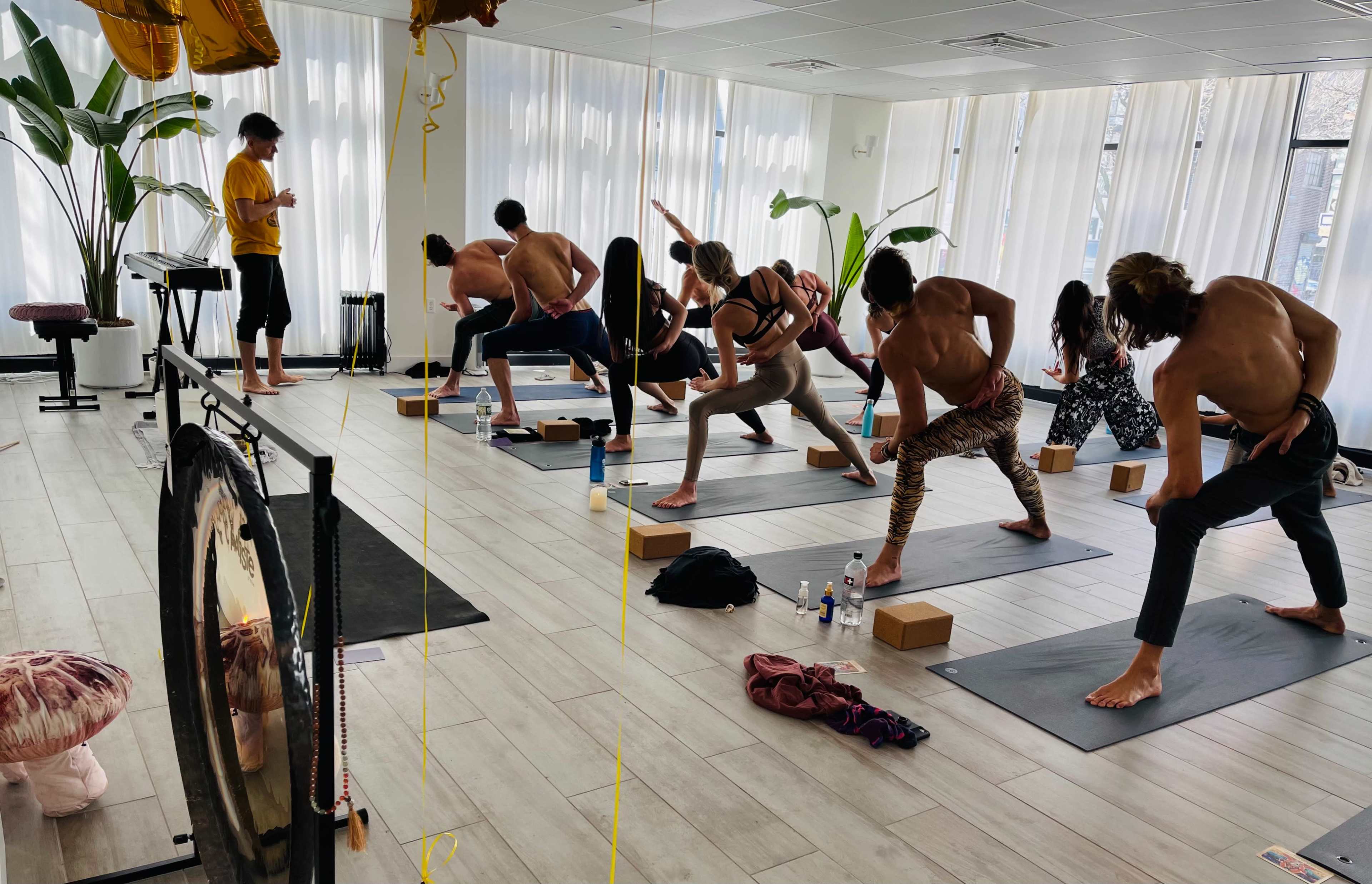 A group of people participates in a yoga class in a well-lit studio with plants and various yoga props.