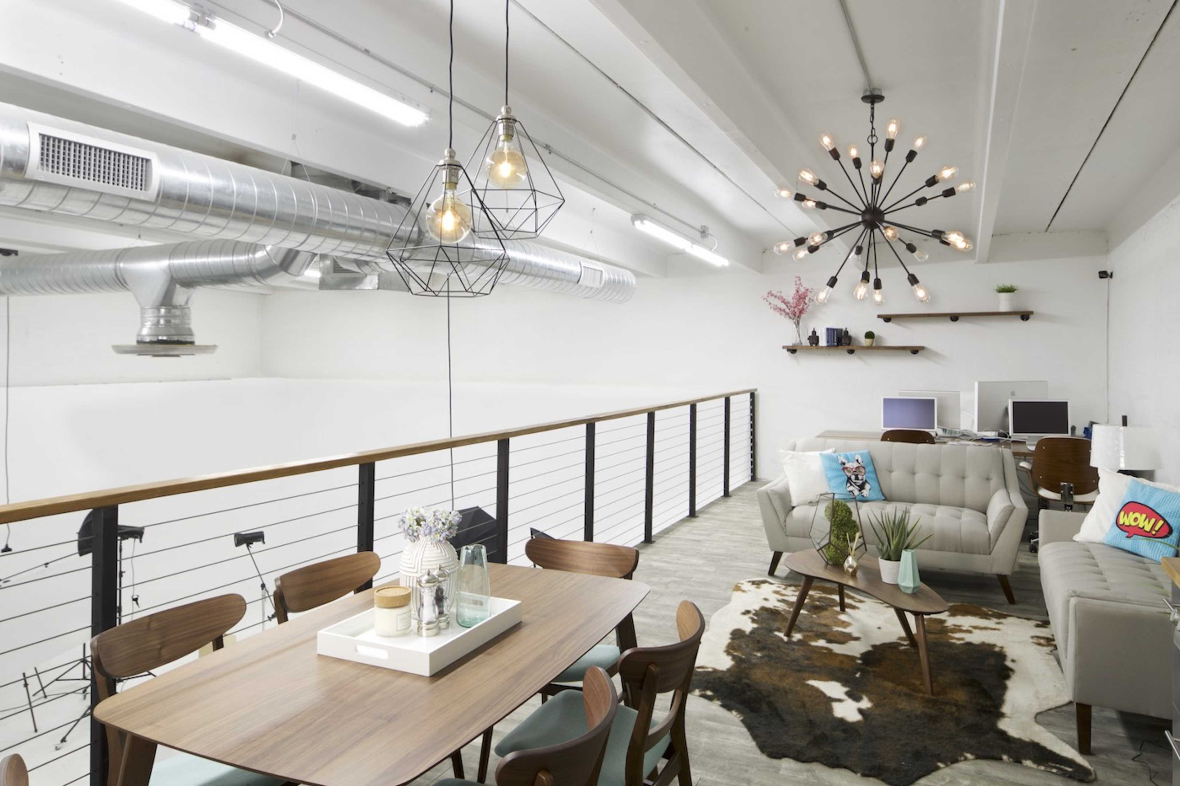 The image shows a modern loft-style interior with a dining table, a decorative sofa arrangement, and industrial lighting fixtures.