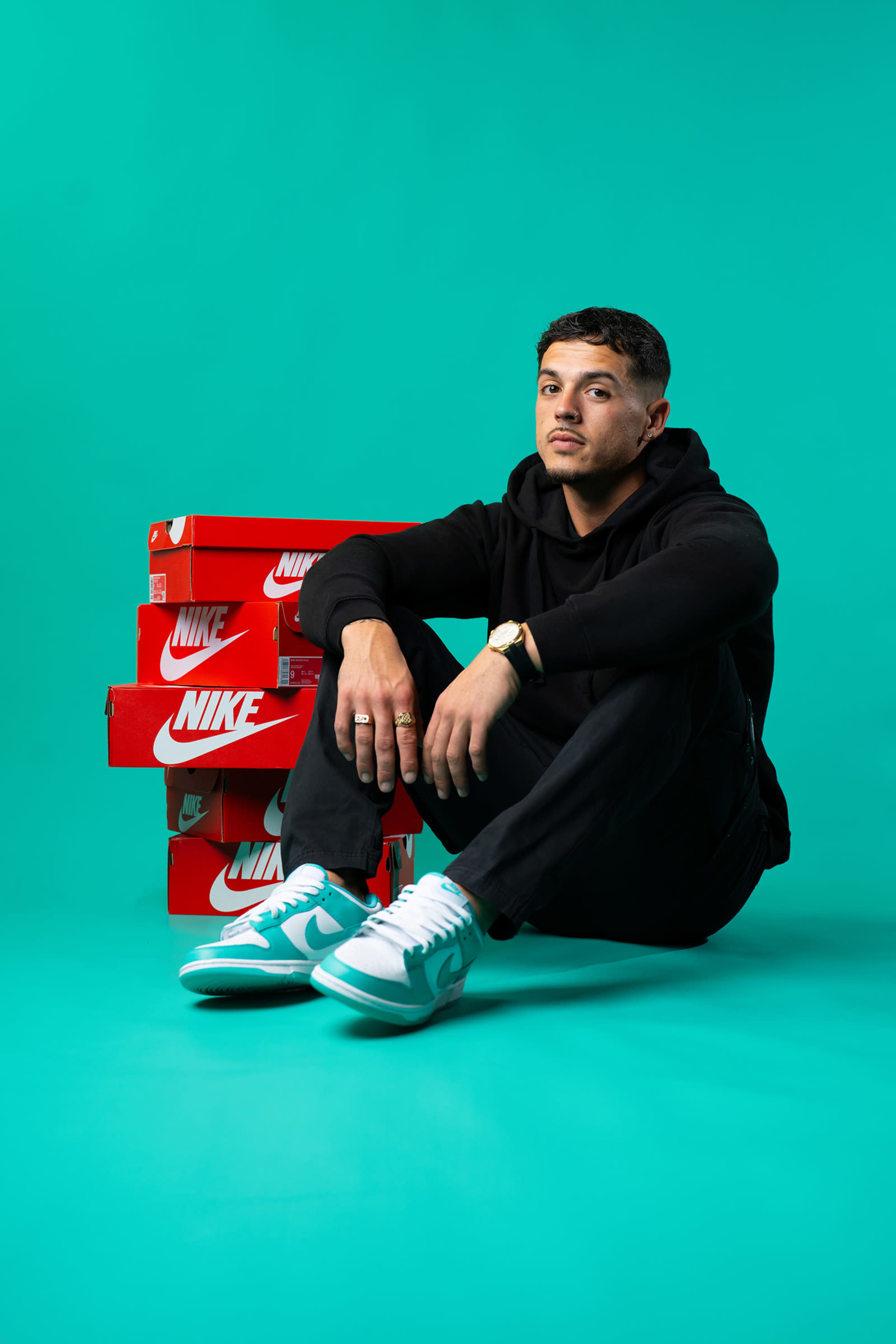 A man sits in front of a stack of red Nike shoeboxes, wearing a black hoodie and light blue sneakers, against a vibrant teal background.