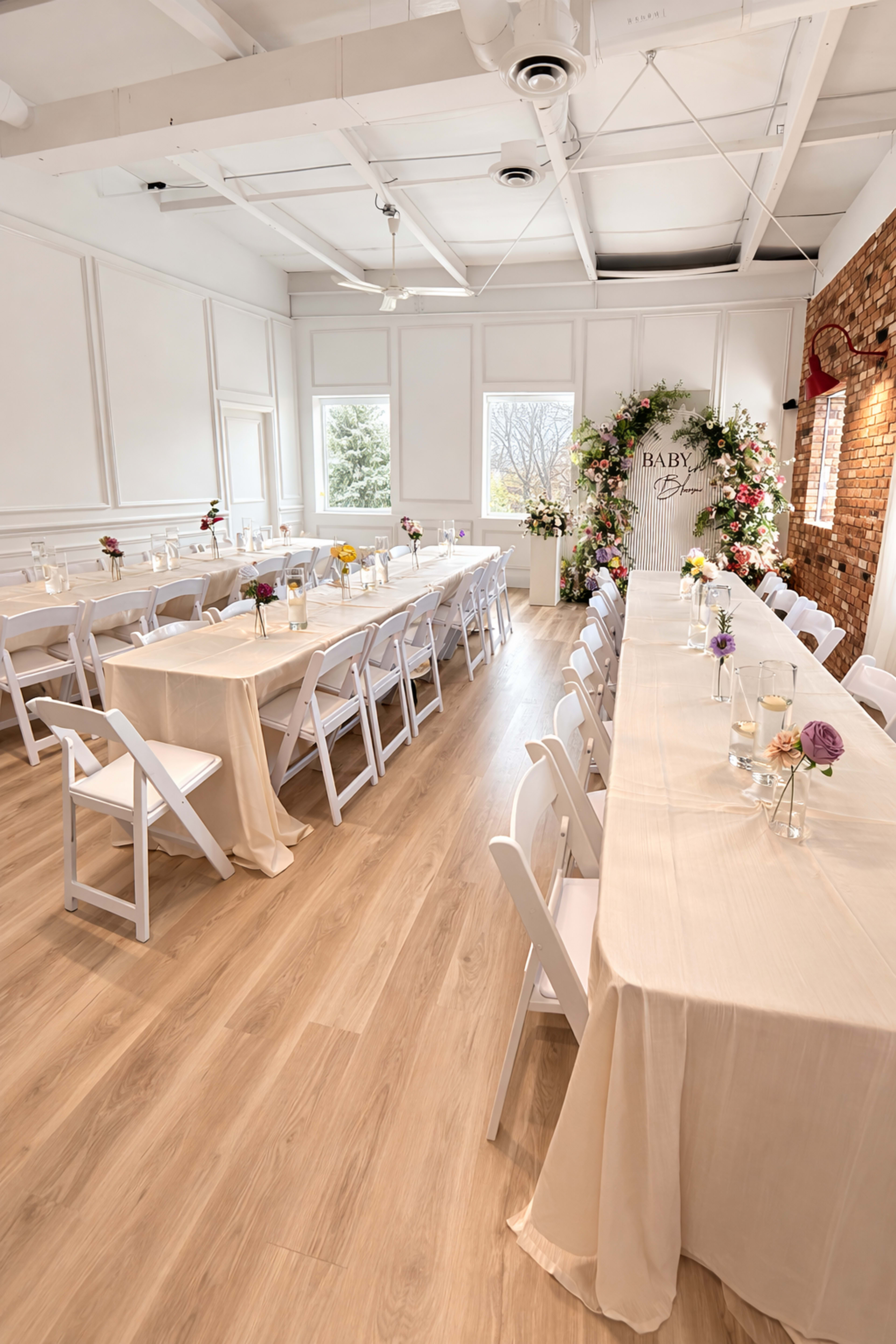 The image shows a decorated indoor event space set up with long tables covered in beige cloth, surrounded by white folding chairs, and featuring a floral backdrop with a "Baby" sign.