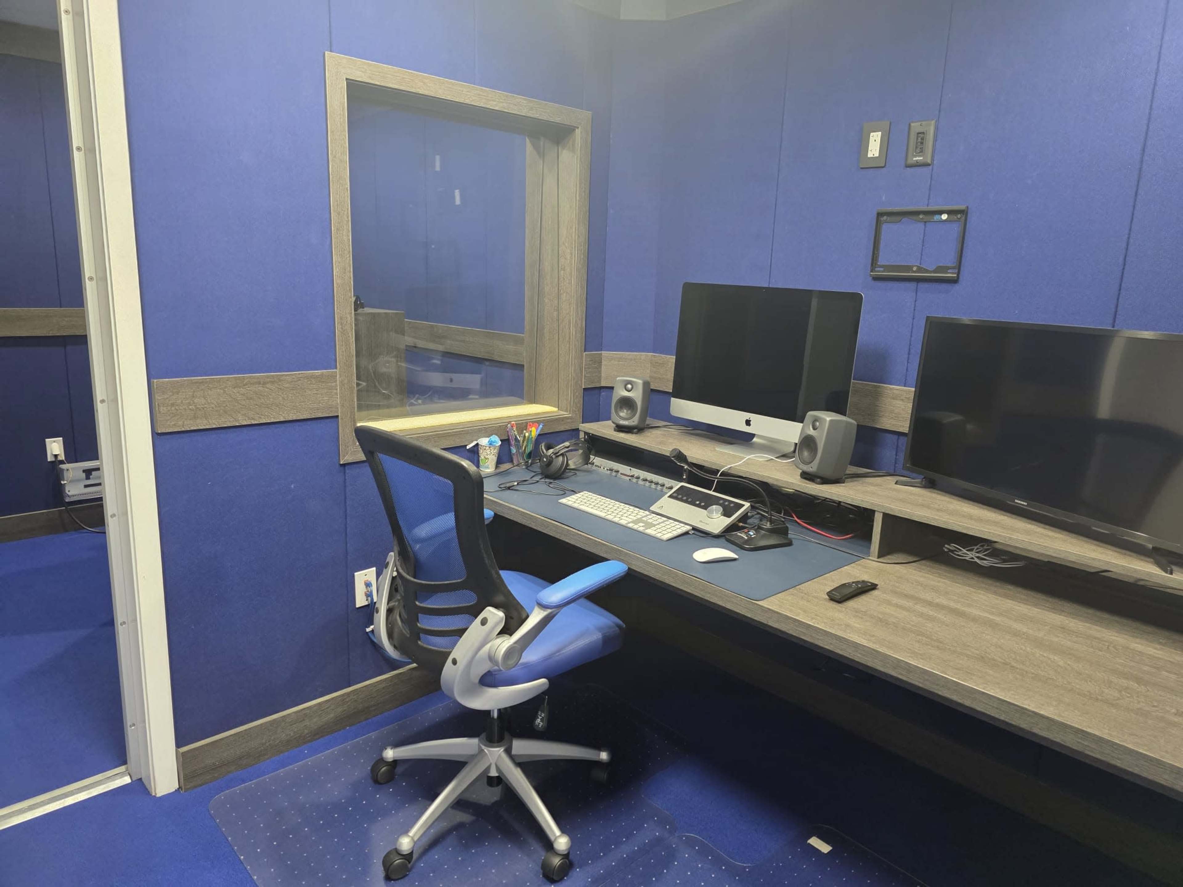 The image shows a small office or studio space with blue walls, a large desk featuring two computer monitors and an iMac, and a rolling office chair.