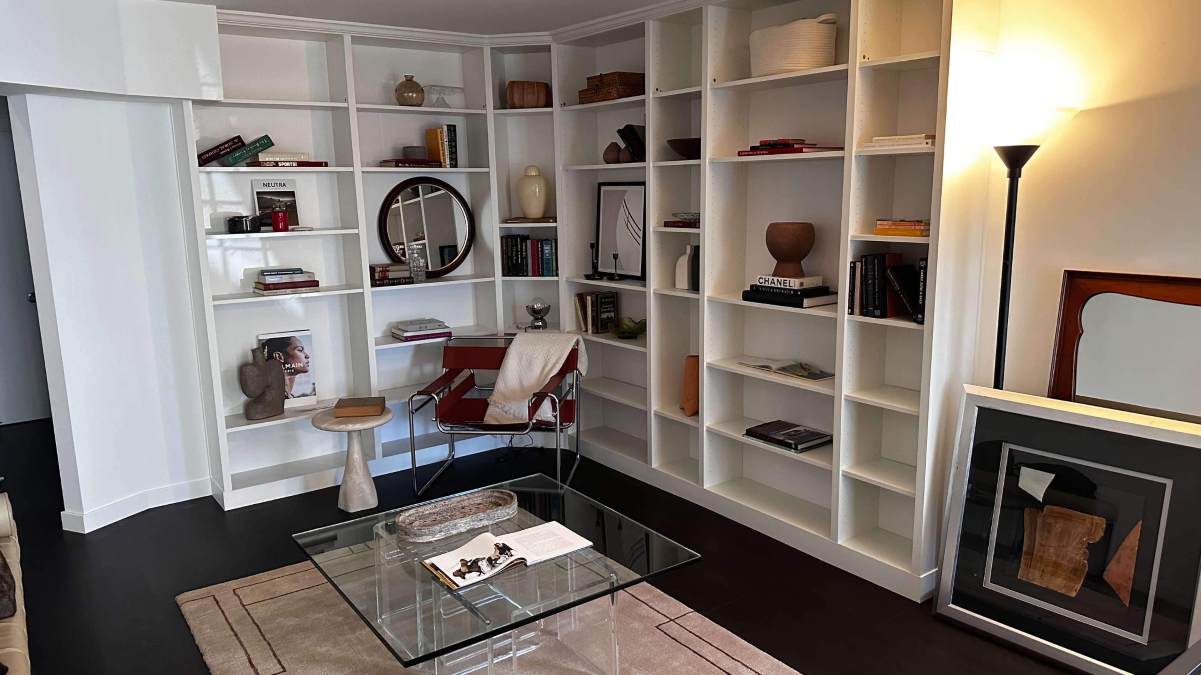 A living room features a glass coffee table, a small side table, a red chair, and built-in shelves filled with books and decorative items.