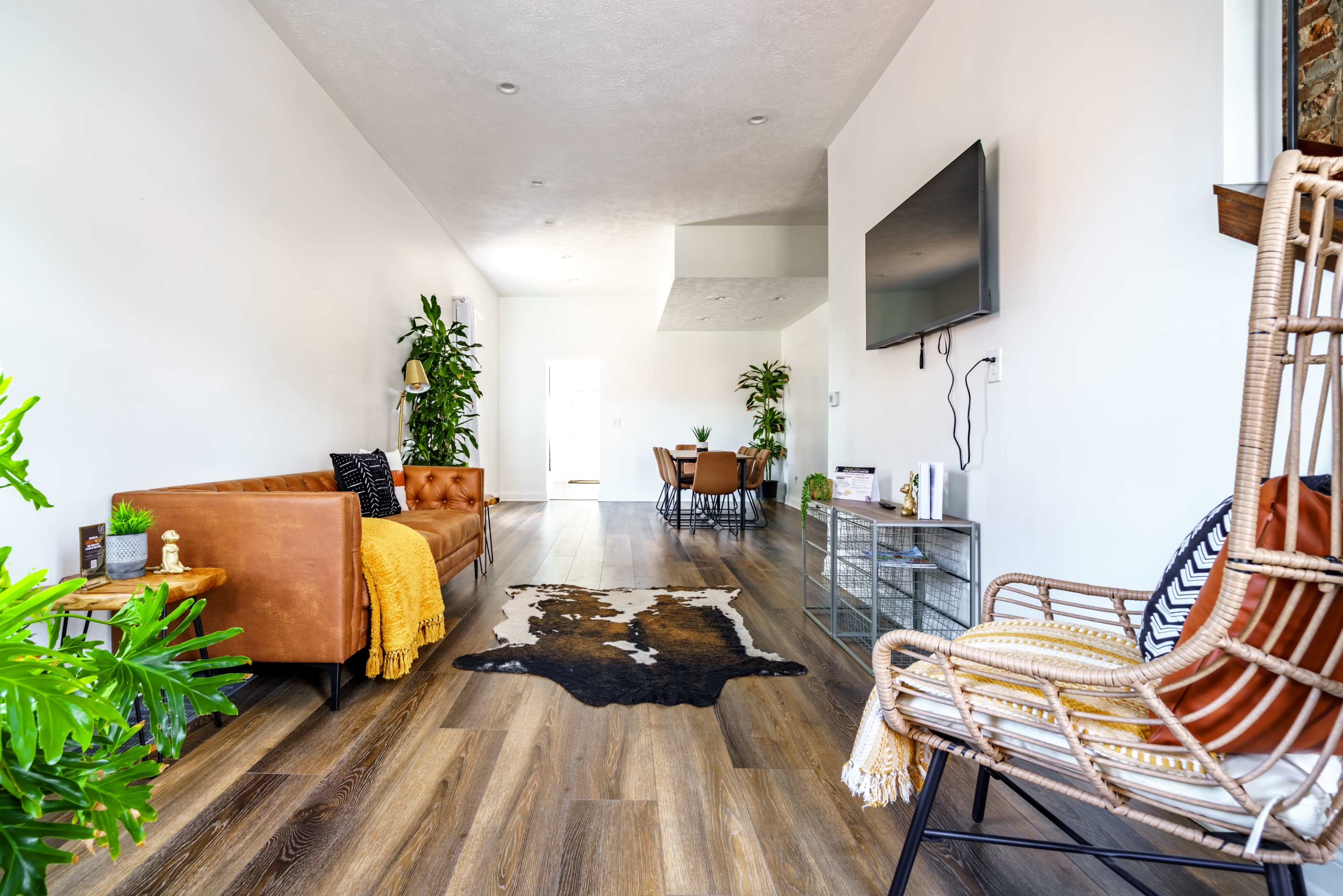 The image shows a modern, open living space featuring a brown leather sofa, a decorative cowhide rug on wooden flooring, a TV mounted on the wall, and a dining area visible in the background.