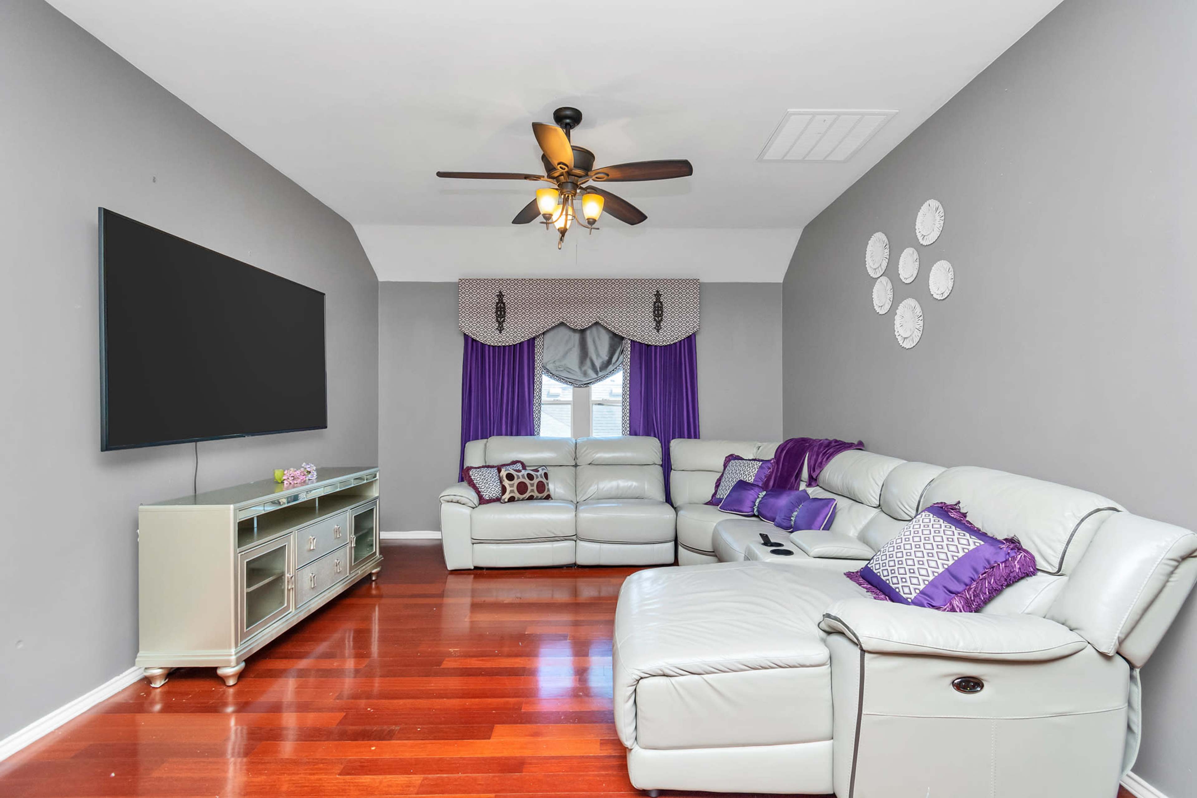 A modern living room features a large flat-screen TV, a light-colored leather sectional sofa, and purple accents on the curtains and throw pillows against gray walls.