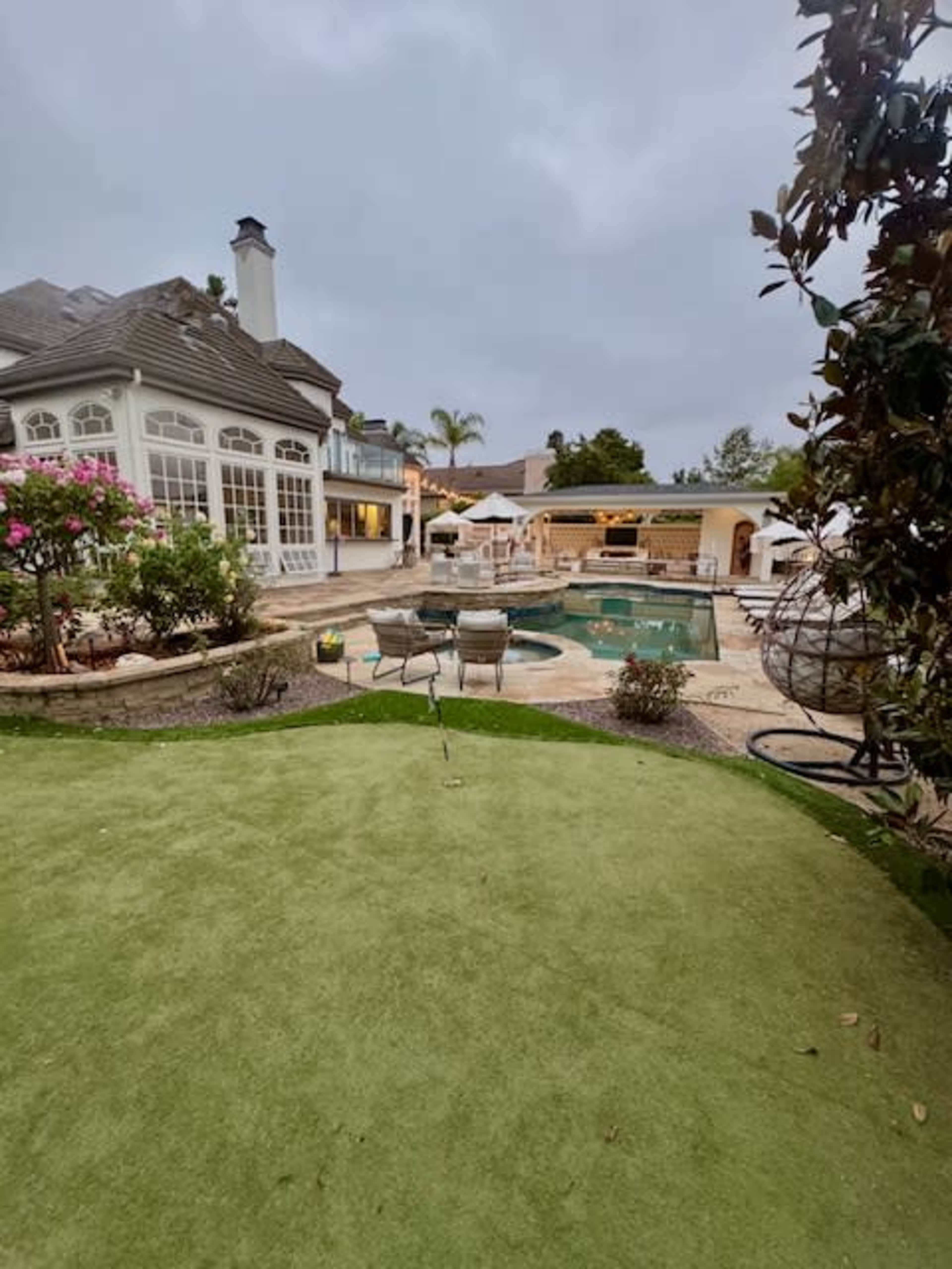 The image shows a landscaped backyard with a swimming pool, spa, and outdoor seating areas adjacent to a house, all under a cloudy sky.