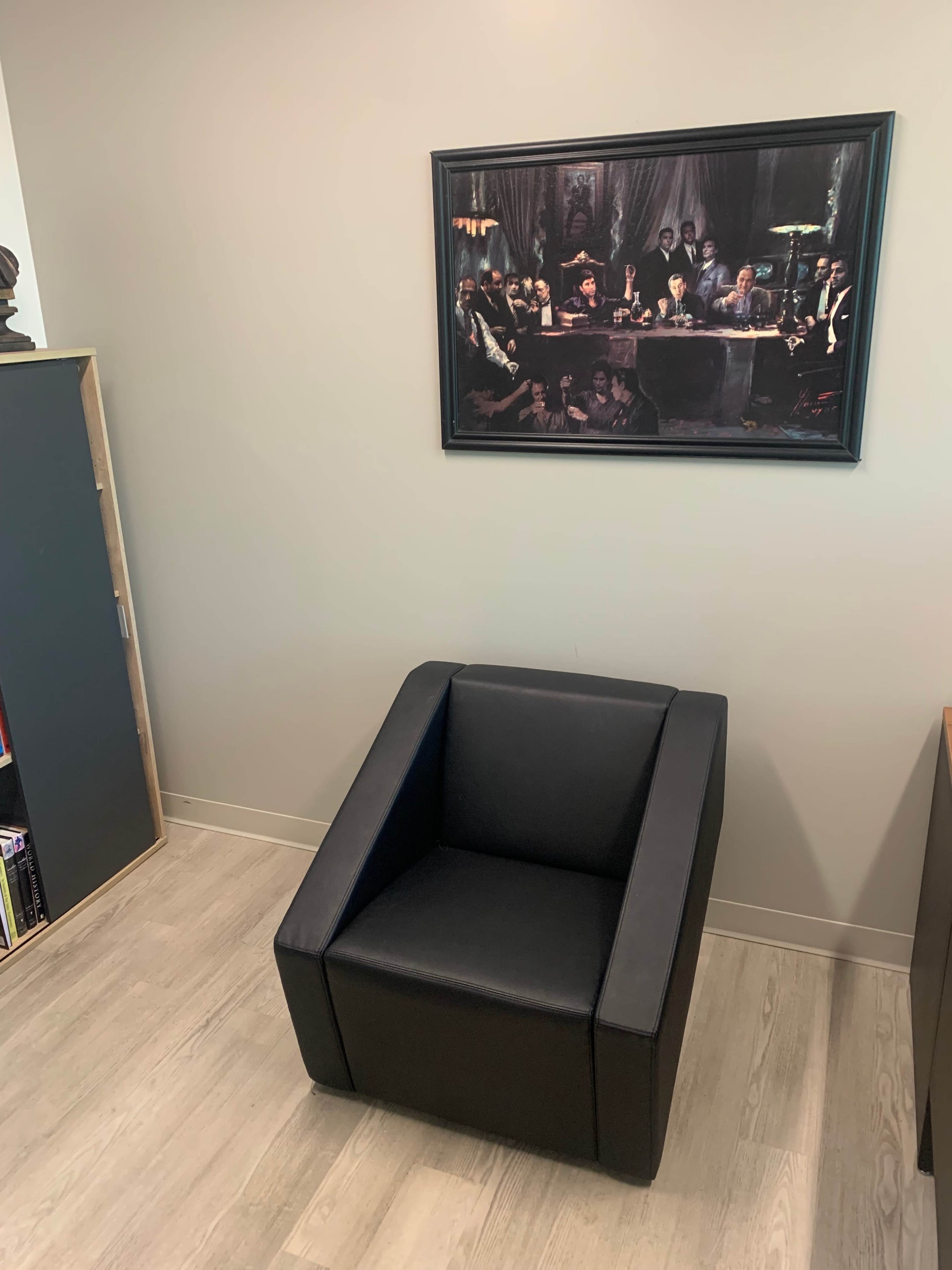 A black armchair is positioned in a corner beneath a framed painting that depicts a group of people at a table.