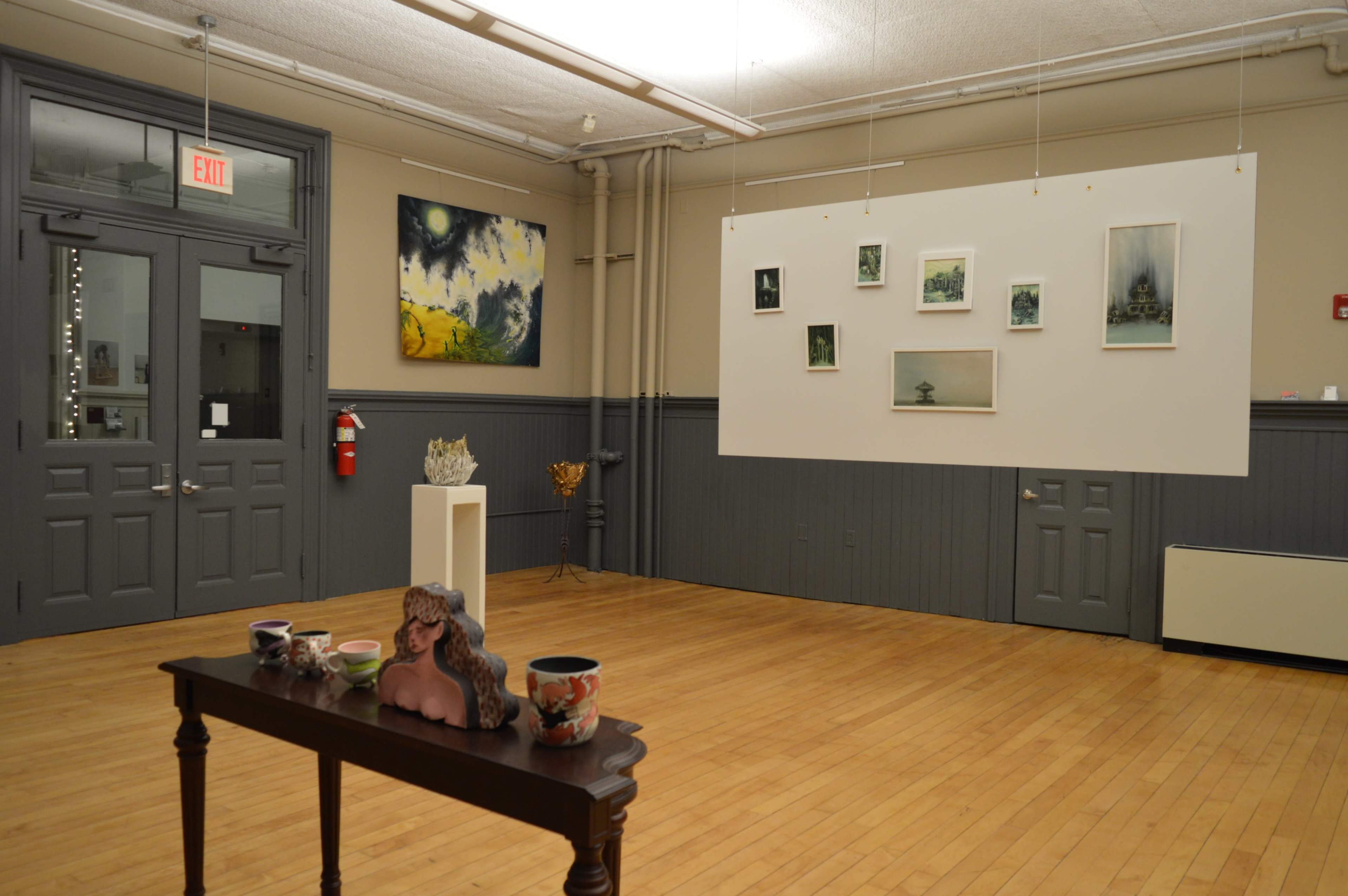 An art gallery featuring a table with ceramic pieces and a bust sculpture, alongside a wall displaying framed artwork.