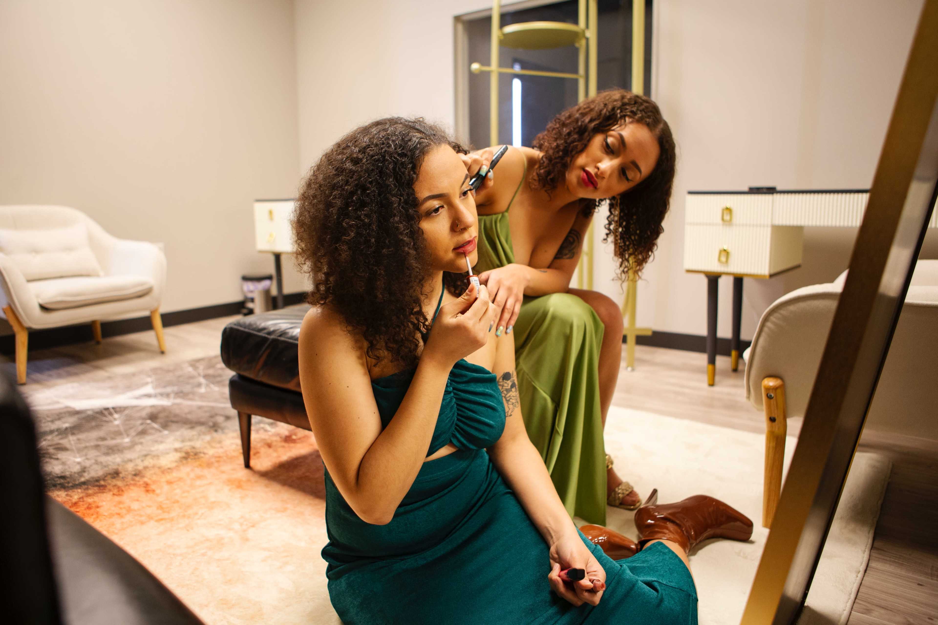 Two women with curly hair are applying makeup in front of a mirror in a modern, well-lit room.