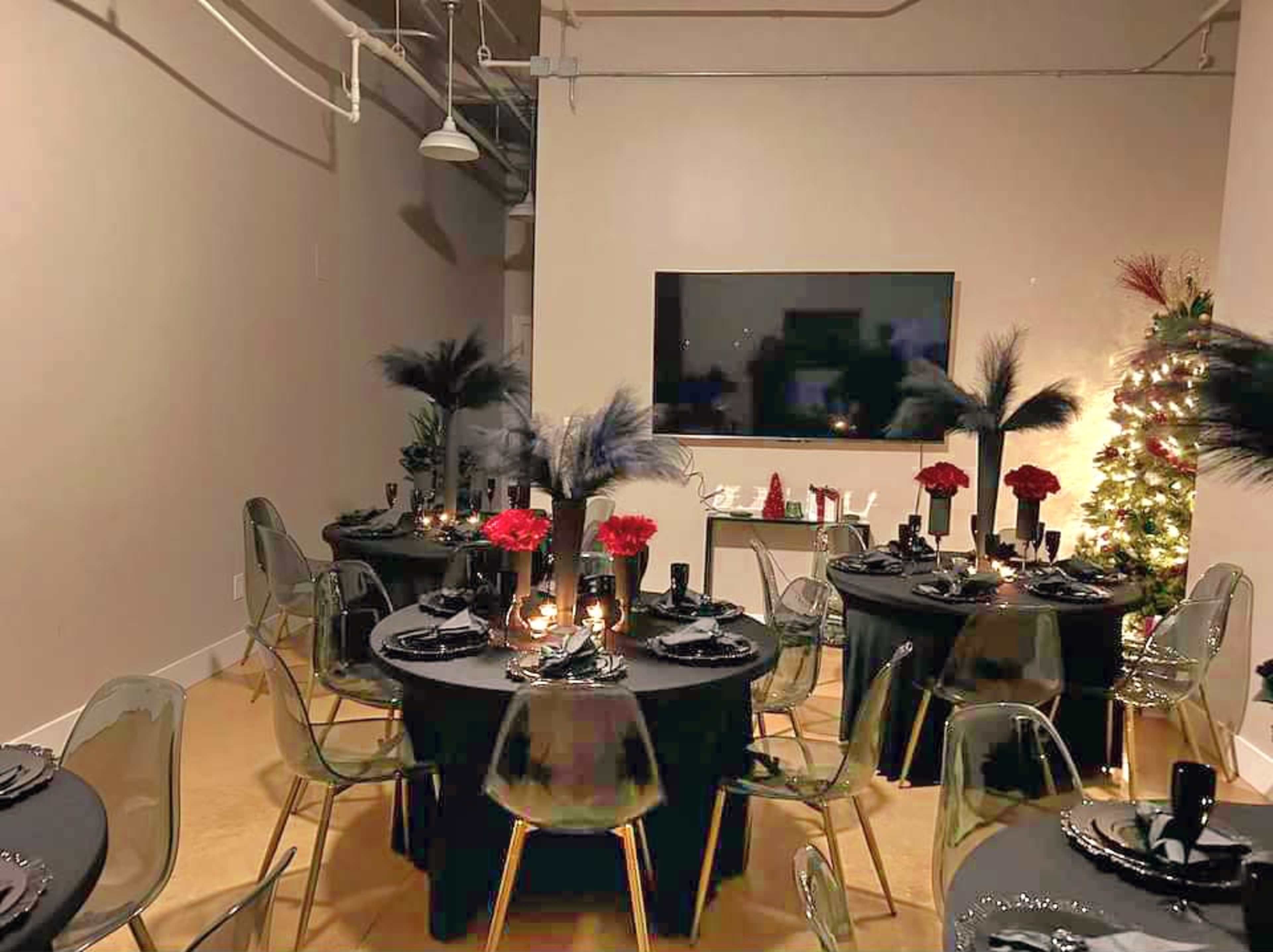 A dining room is set up with black tablecloths, clear chairs, red floral centerpieces, and a decorated Christmas tree in the corner.
