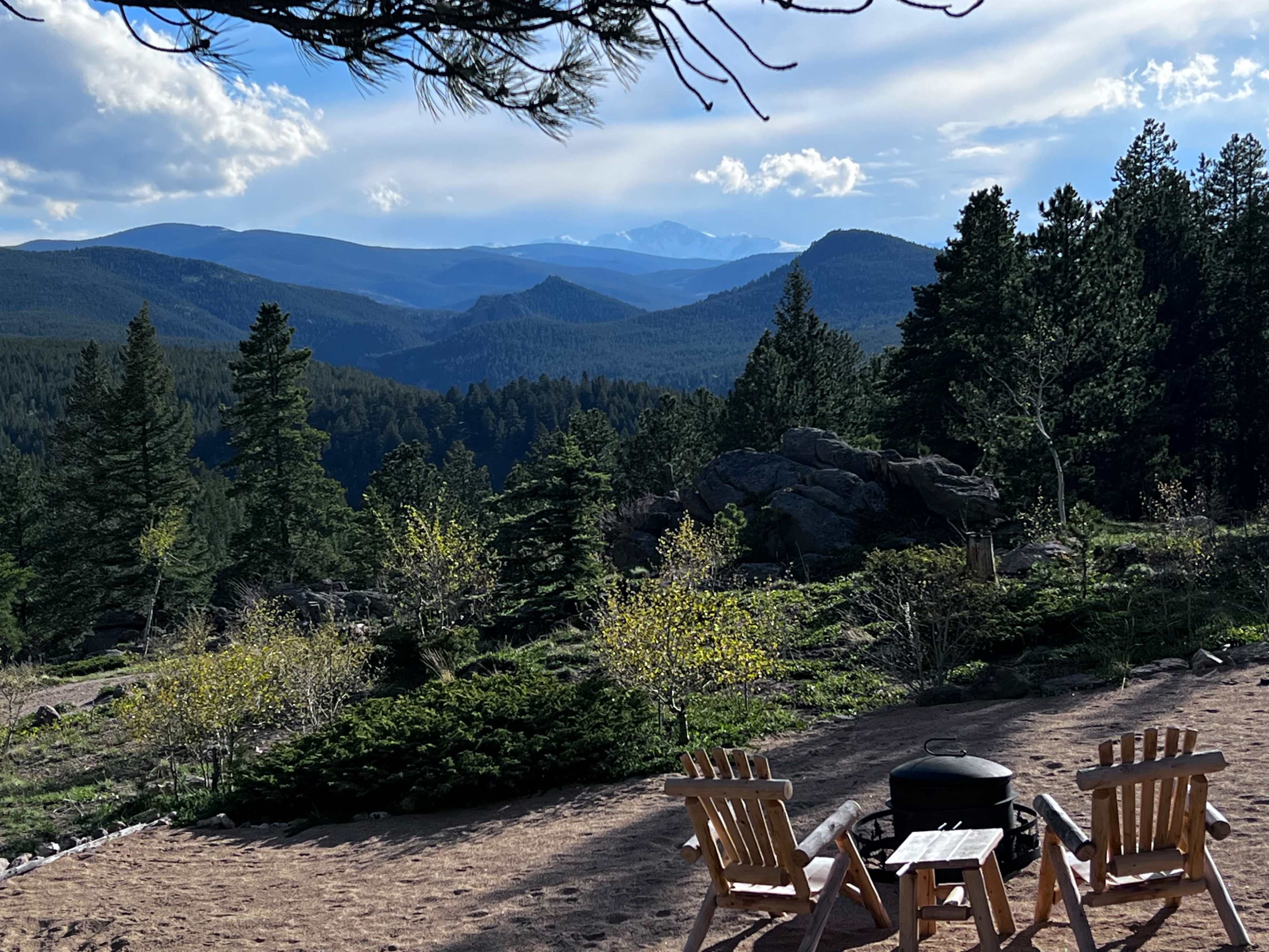 Two wooden chairs face a vast mountainous landscape dotted with trees and distant peaks under a partly cloudy sky.