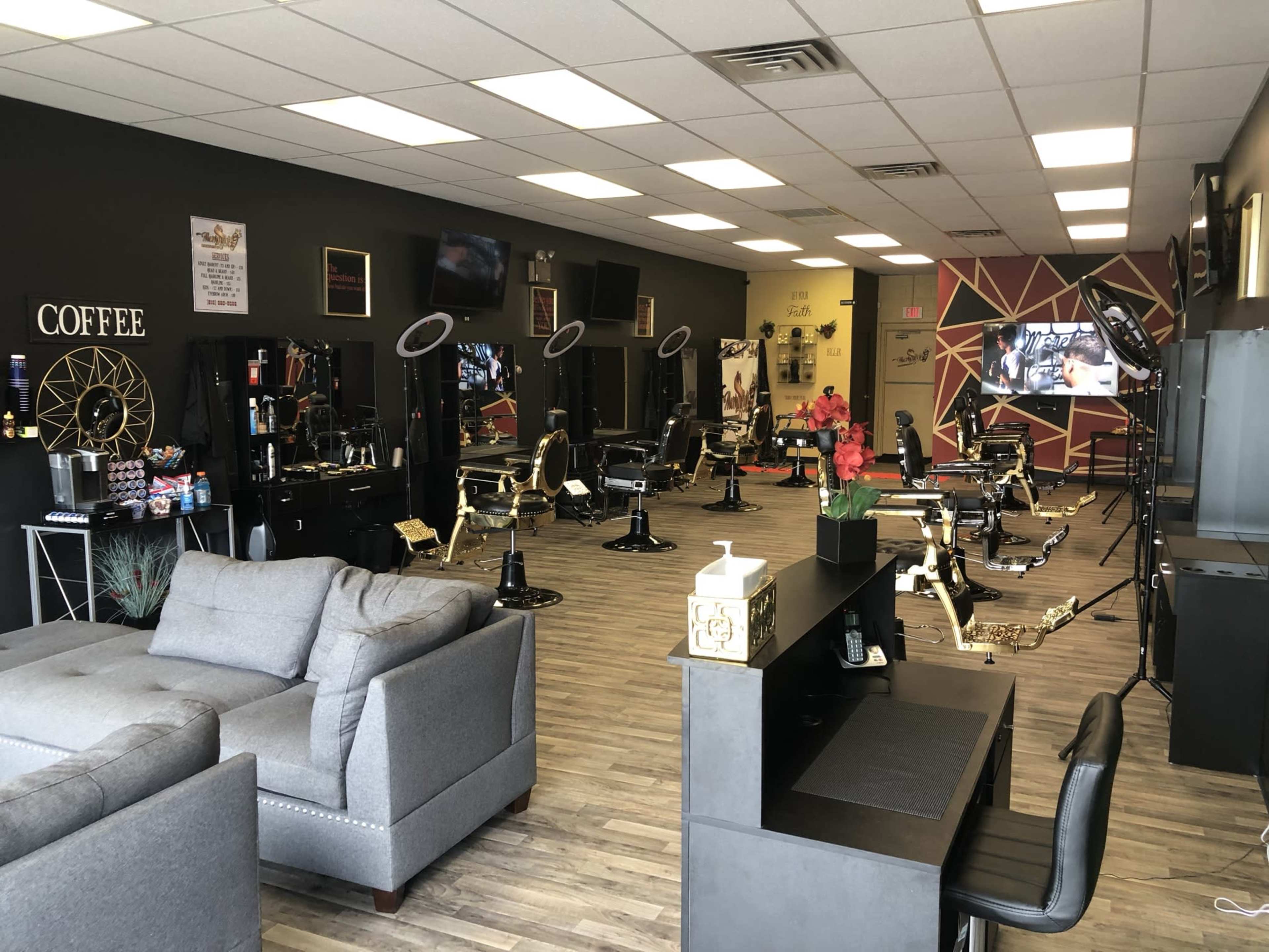 A spacious barbershop with several styling stations, comfortable seating, and a decorative wall featuring various barber tools and a television screen.