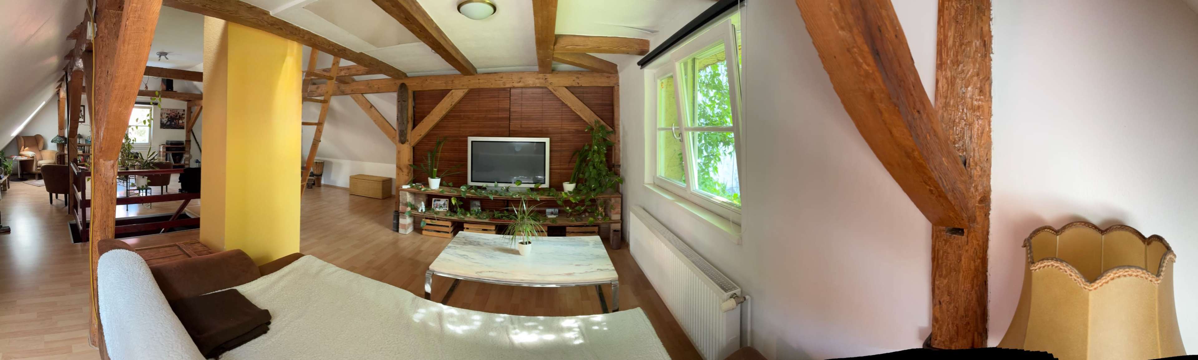 The image shows a spacious room with wooden beams, a large TV on a cabinet, and a coffee table surrounded by light-colored furnishings.