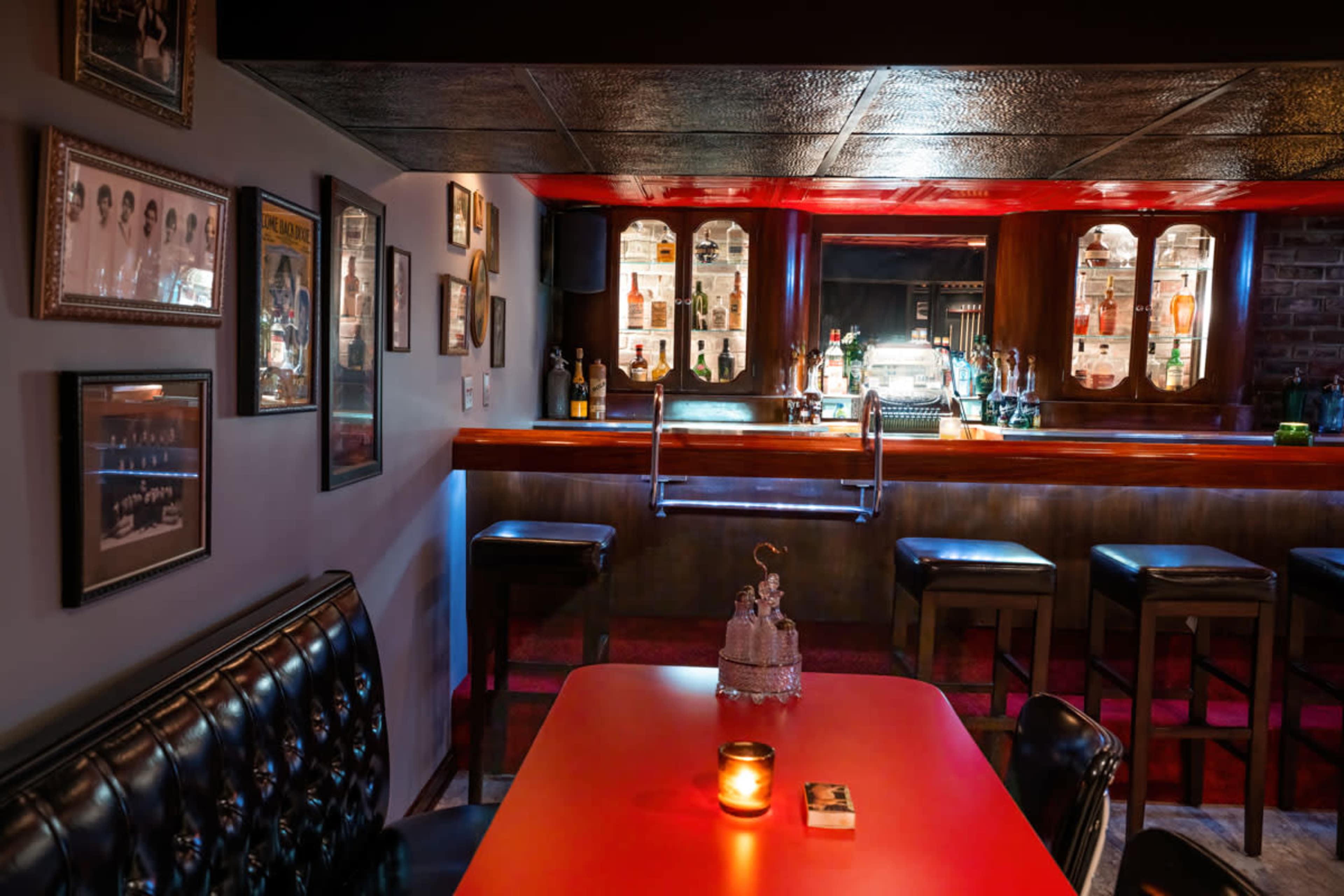 A dimly lit bar area with a polished counter, high stools, and framed photographs on the walls.