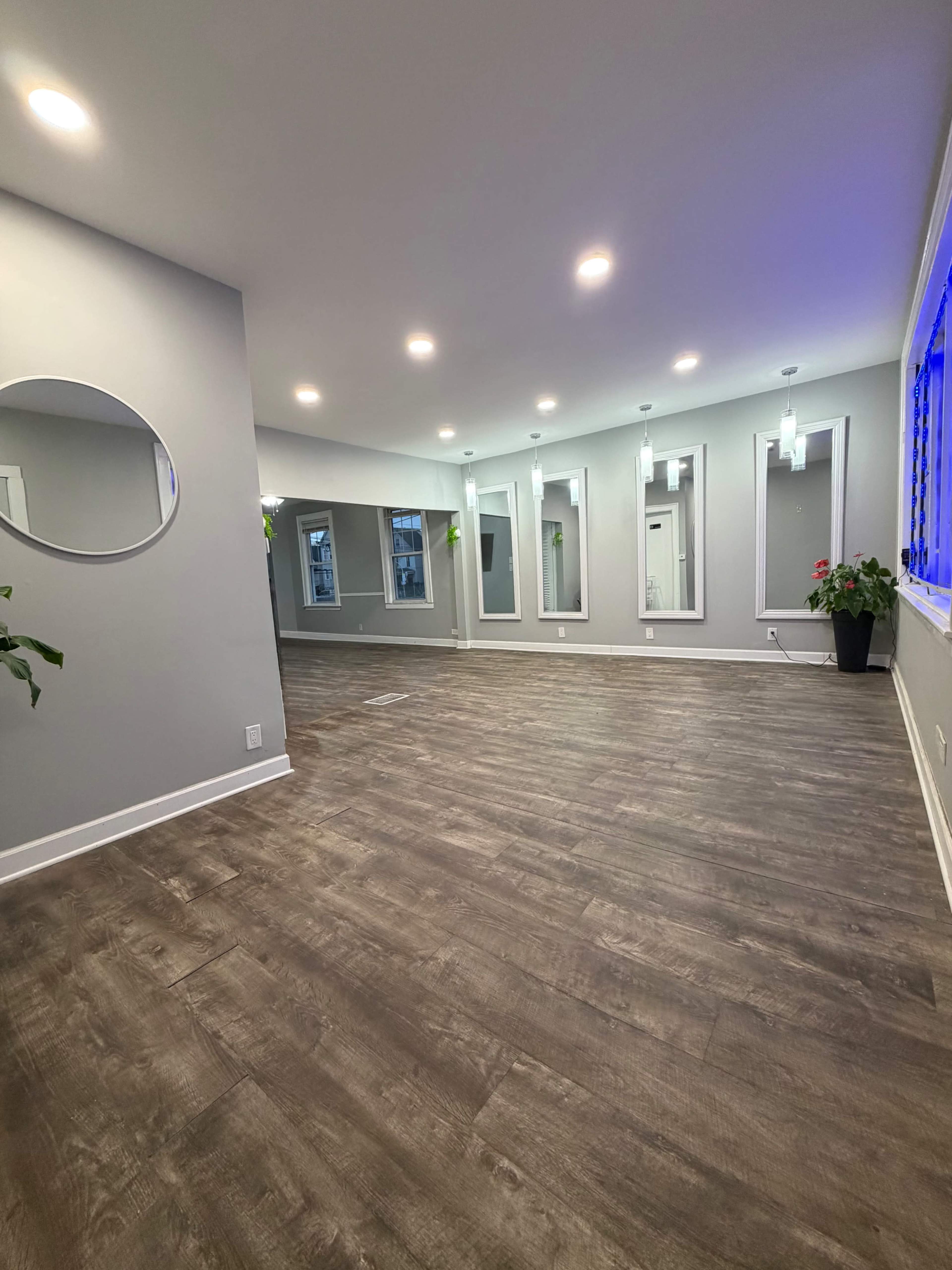 The image shows a spacious interior room with hardwood floors, softly illuminated by recessed lighting, and several framed windows along one wall.