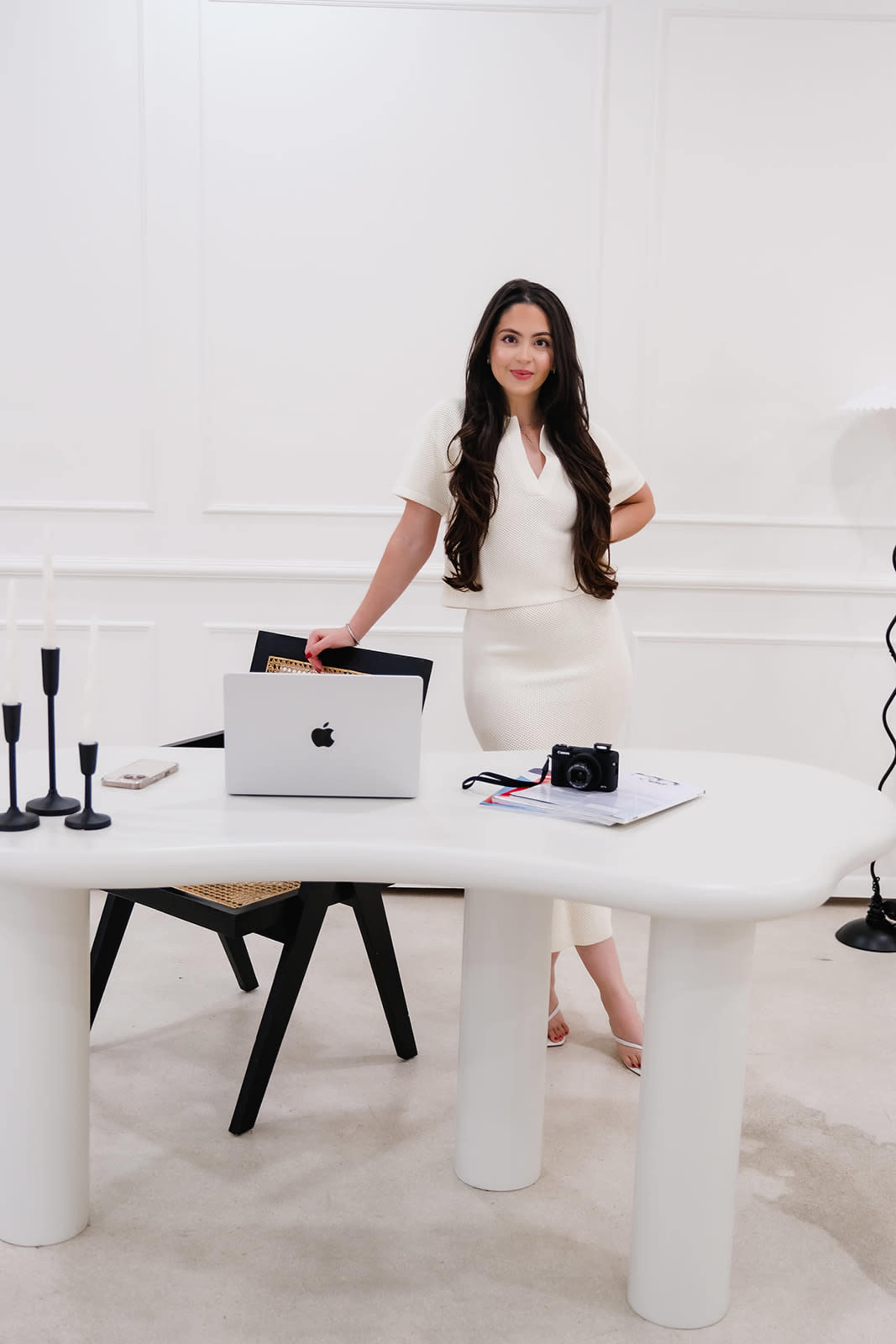 A woman stands beside a modern white table featuring a laptop and a camera, set against a minimalist backdrop.