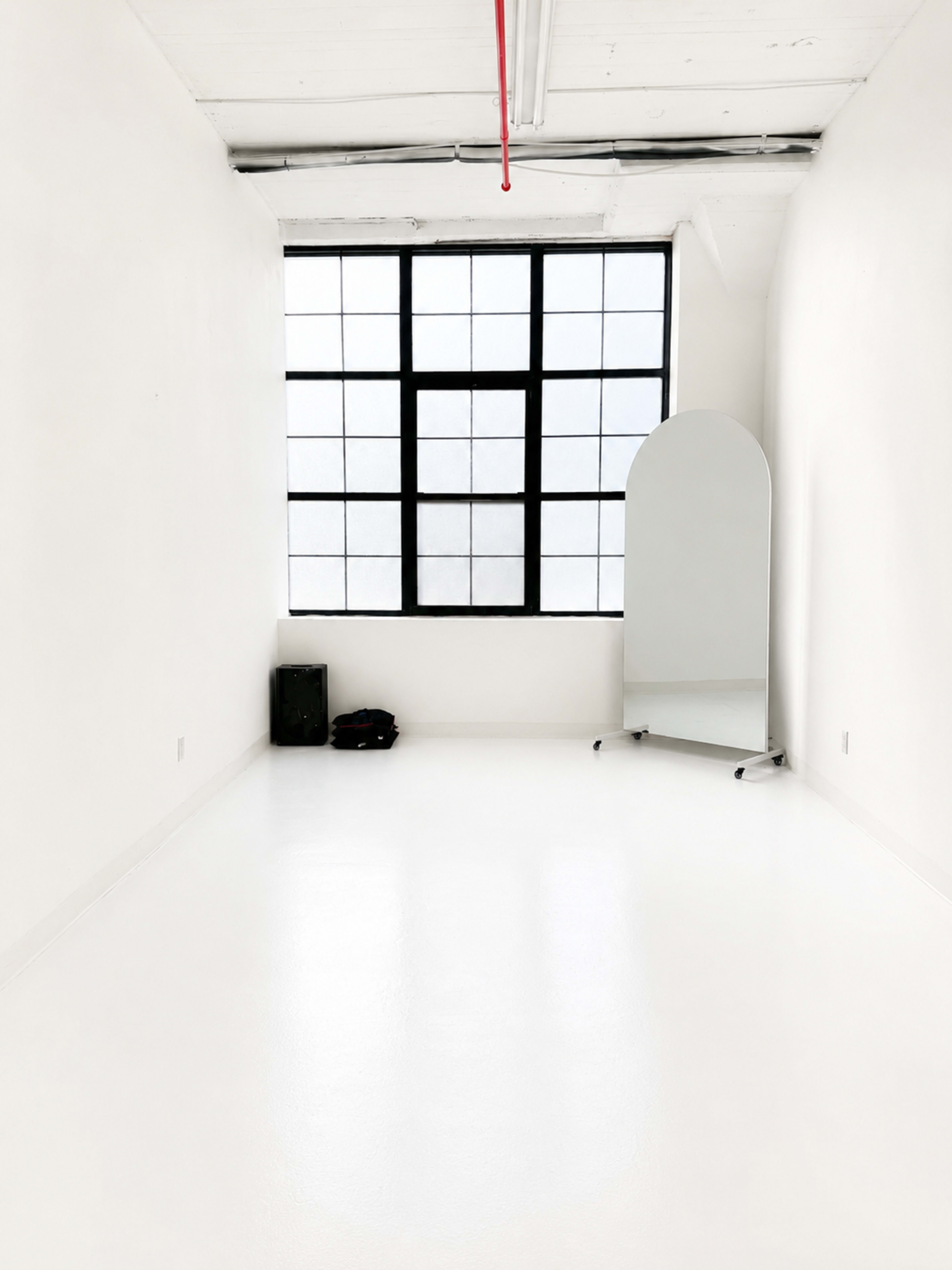 The image shows a minimalistic, empty white room with a large window and a tall mirror on wheels.