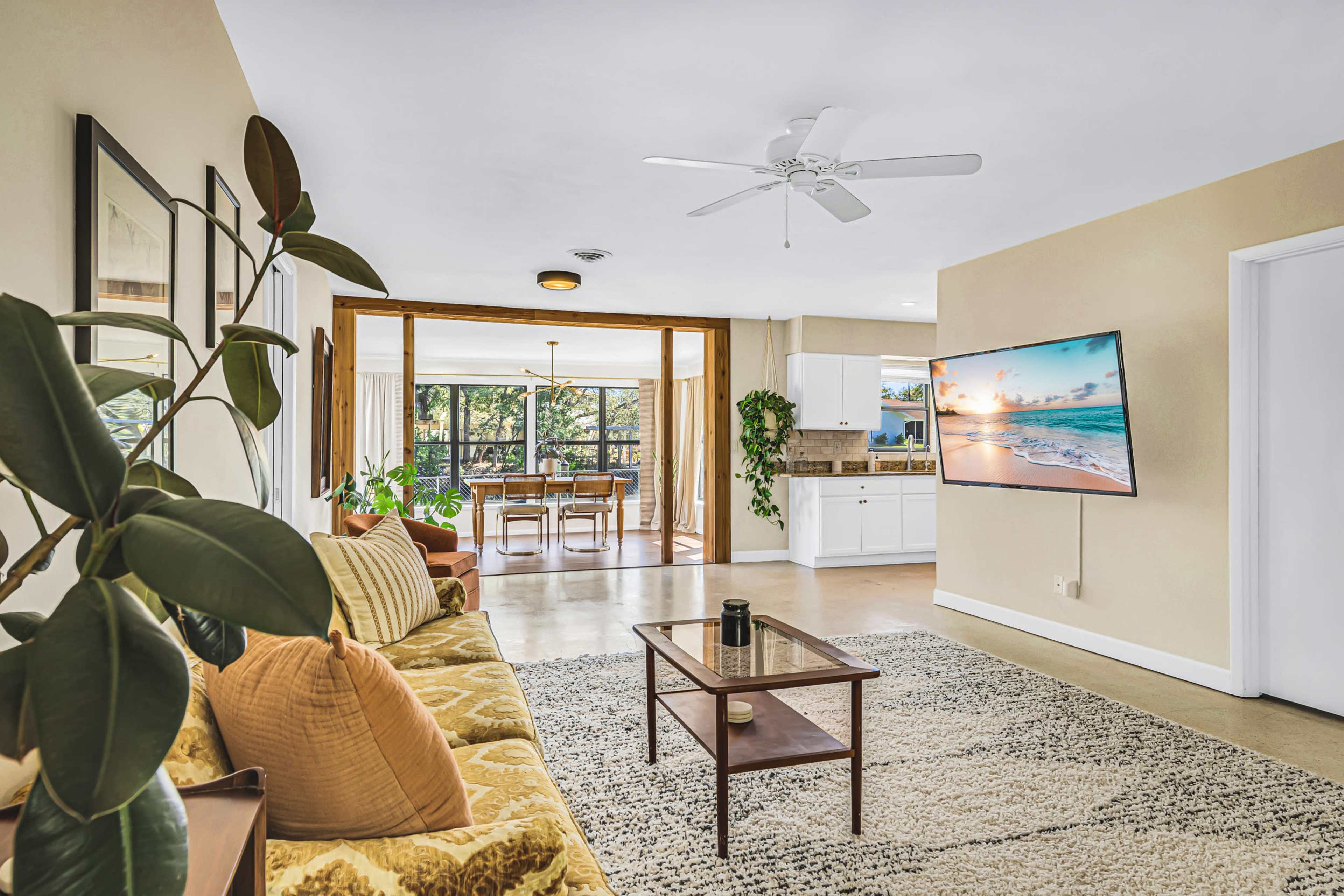A living room features a sofa, a coffee table, and a mounted television, leading to a dining area with large windows that open to a view of greenery.