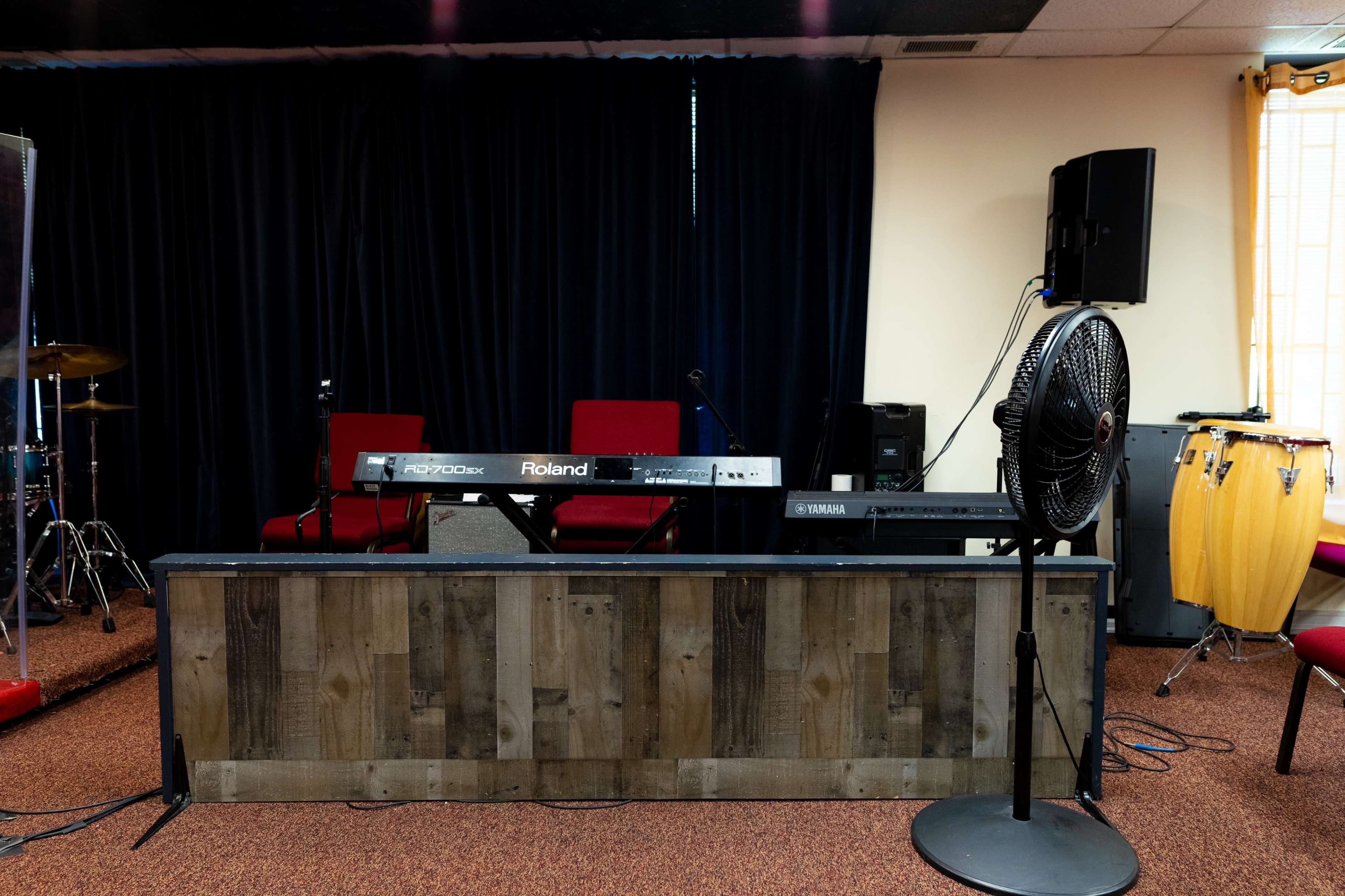 A music performance area features two keyboards, a fan, and various musical instruments against a backdrop of dark curtains and red chairs.