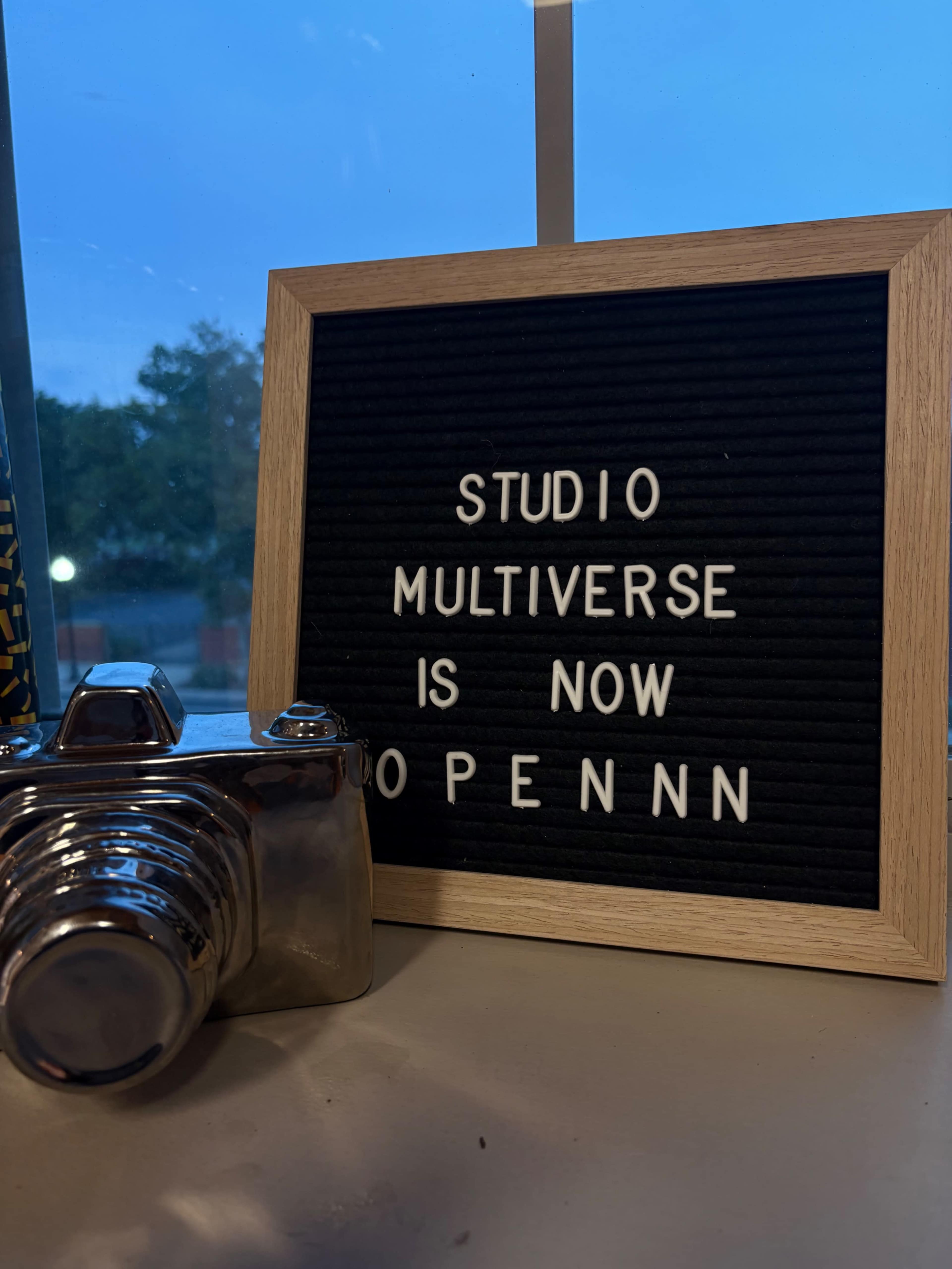 A silver camera sits next to a sign reading "STUDIO MULTIVERSE IS NOW OPENNN" against a blue-lit background.