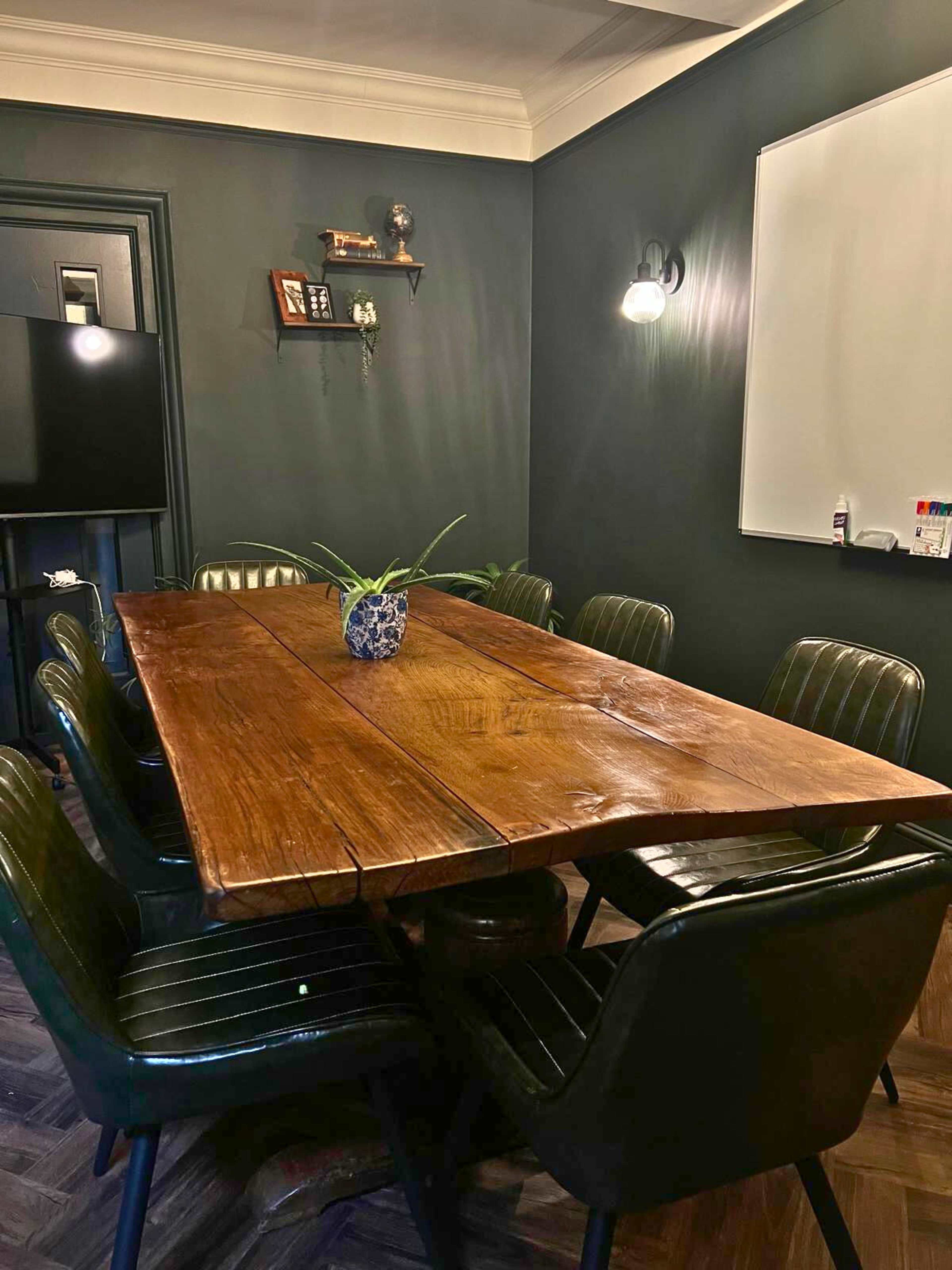 A long wooden table is set in a dark-colored room surrounded by eight black chairs, with a plant in the center and a whiteboard on one wall.