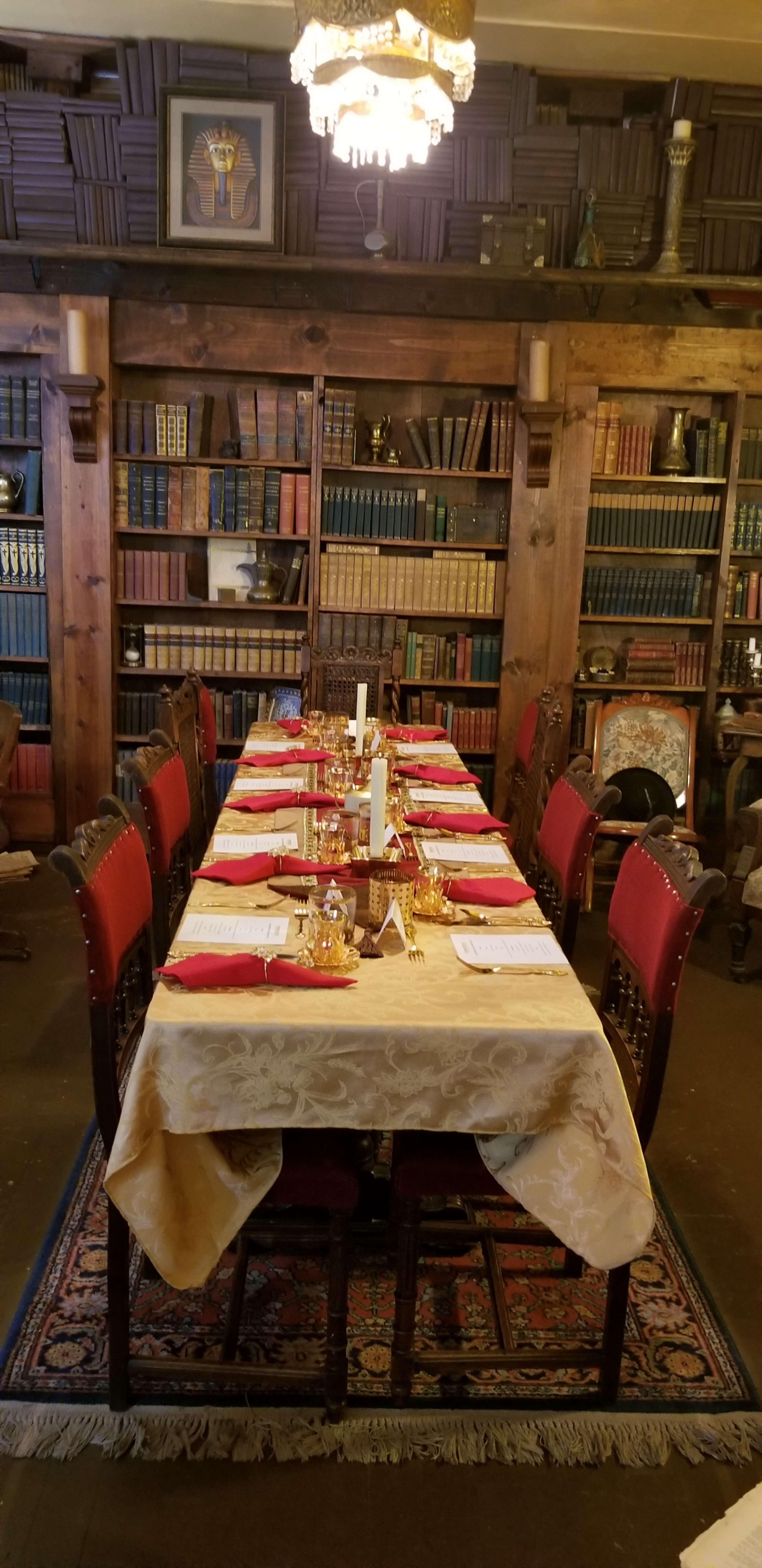 A long dining table is set with red napkins and gold accents in front of wooden bookshelves filled with books.