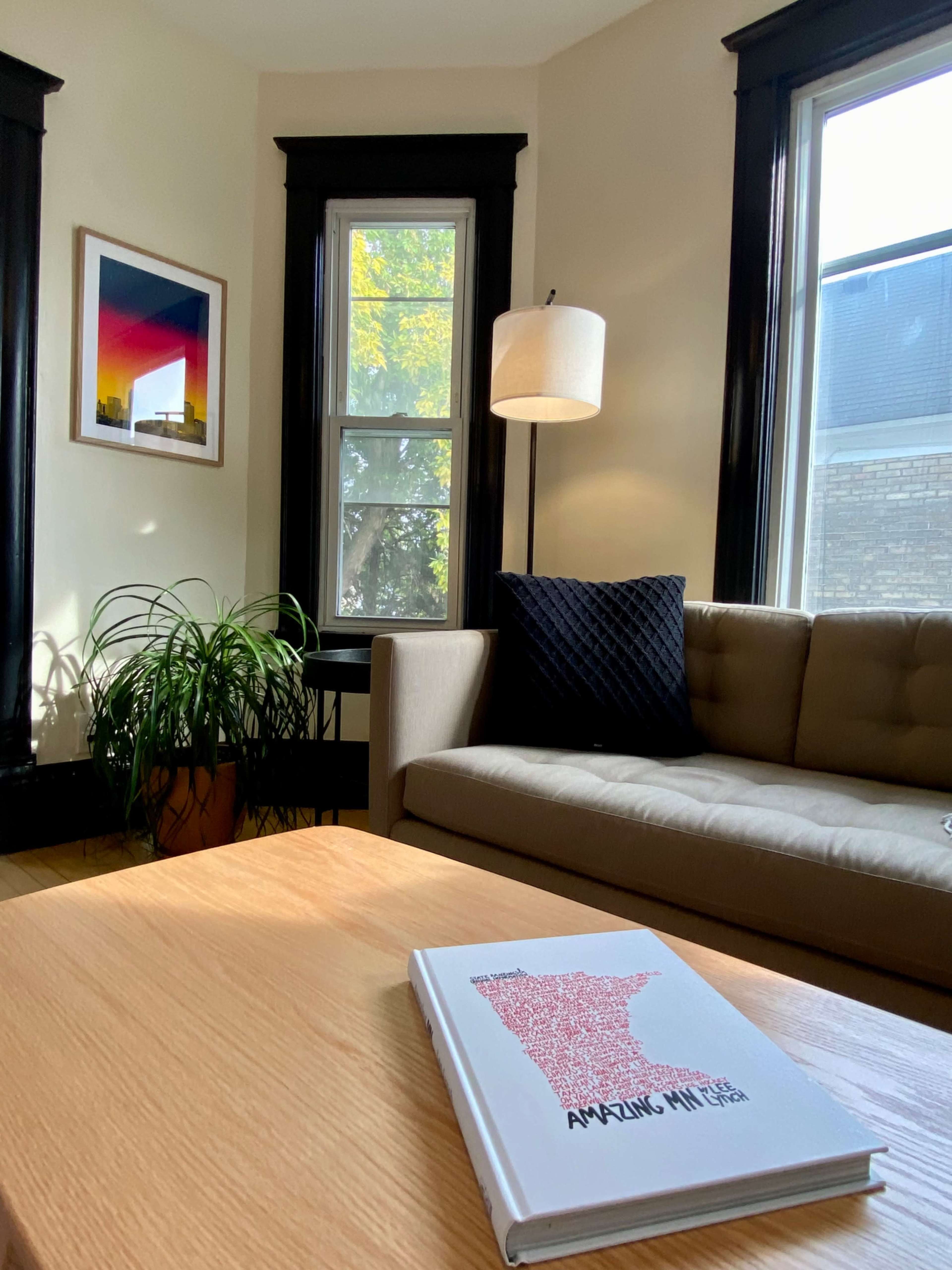 The image shows a cozy living room with a beige sofa, a wooden coffee table, a decorative plant, and large windows framed in black.