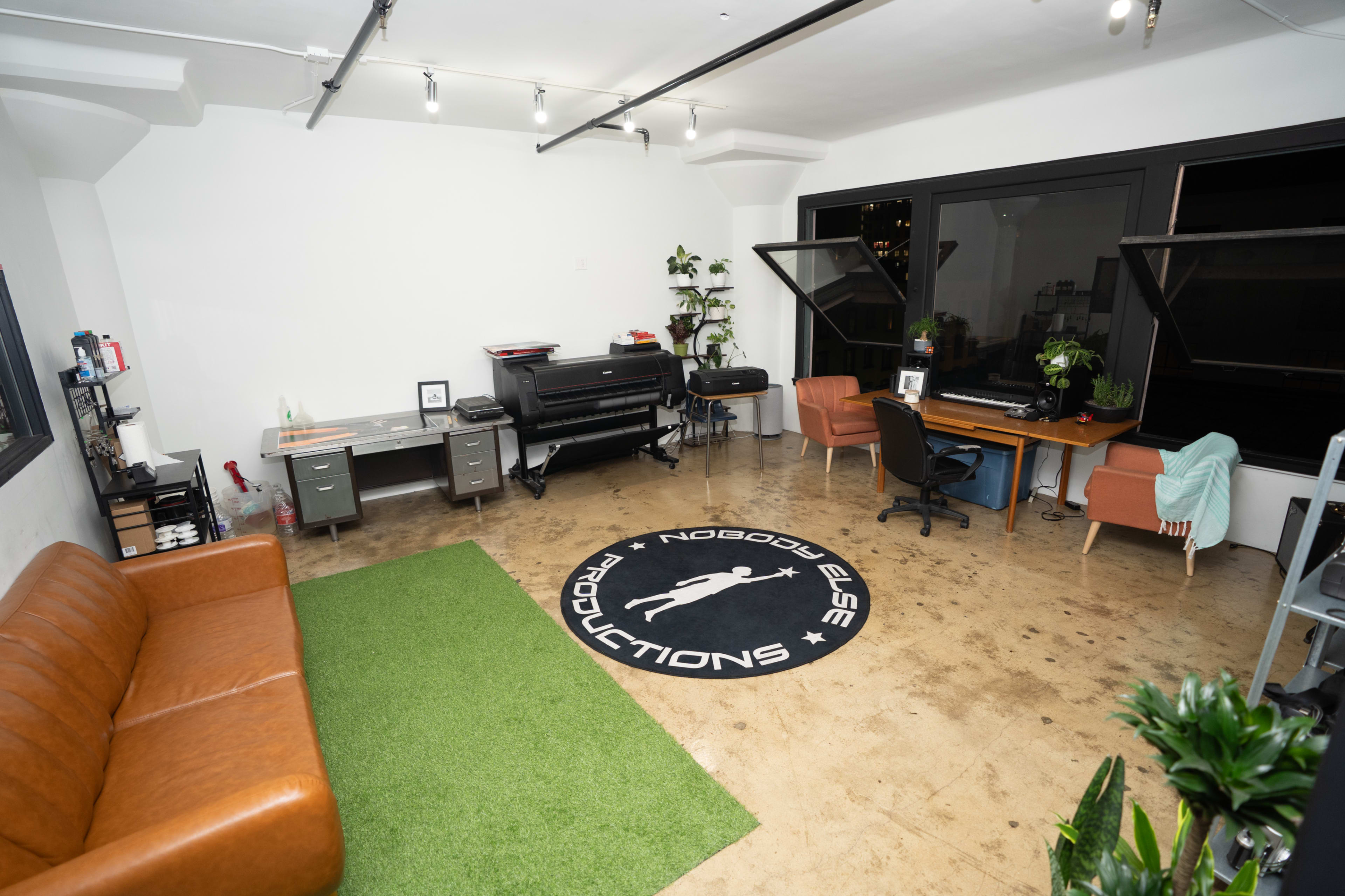 The image shows a spacious office with a brown leather sofa, a green rug, desks with office equipment, and large windows.