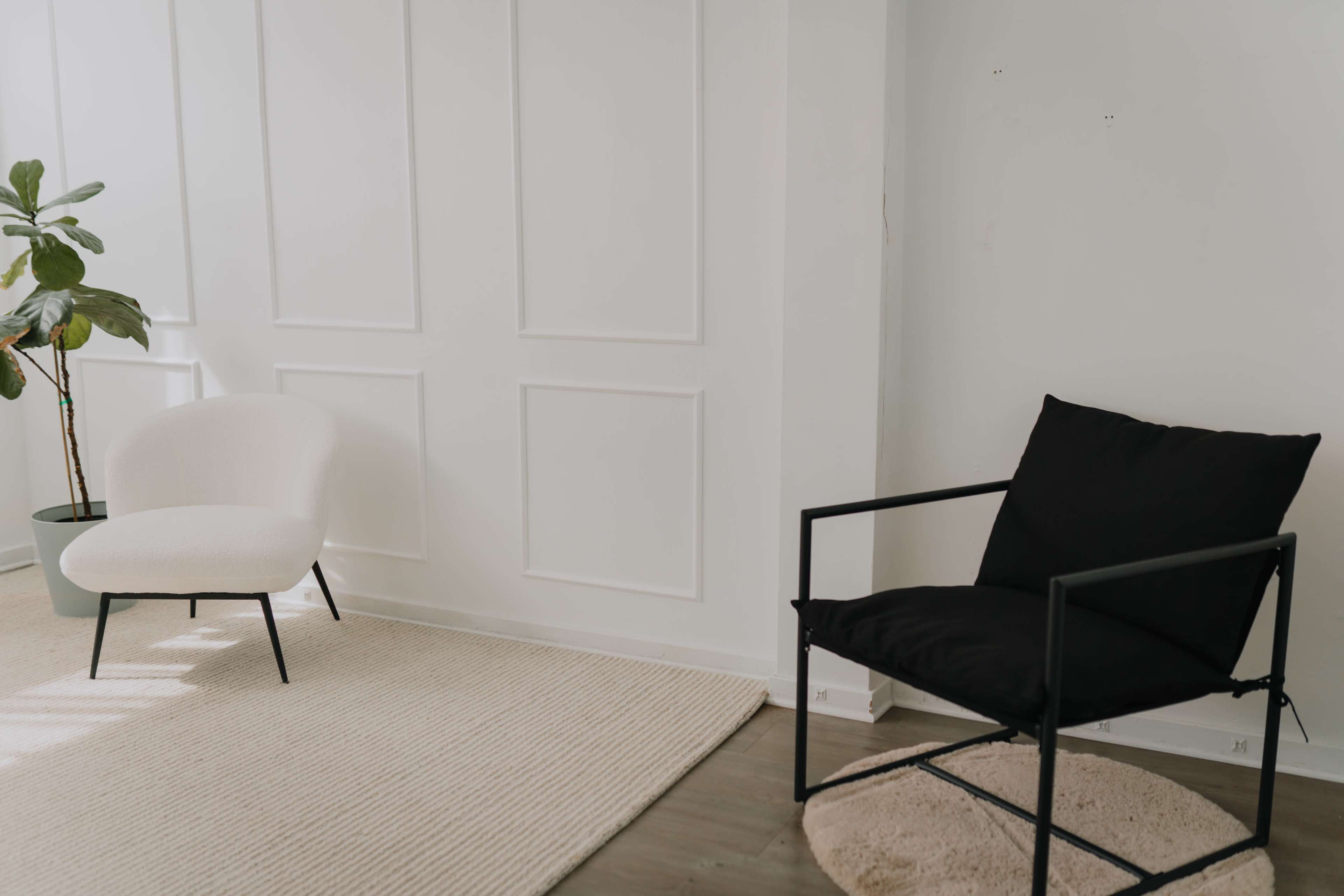 The image shows a minimalist living space with a white chair, a black chair, and a textured area rug on wooden flooring.