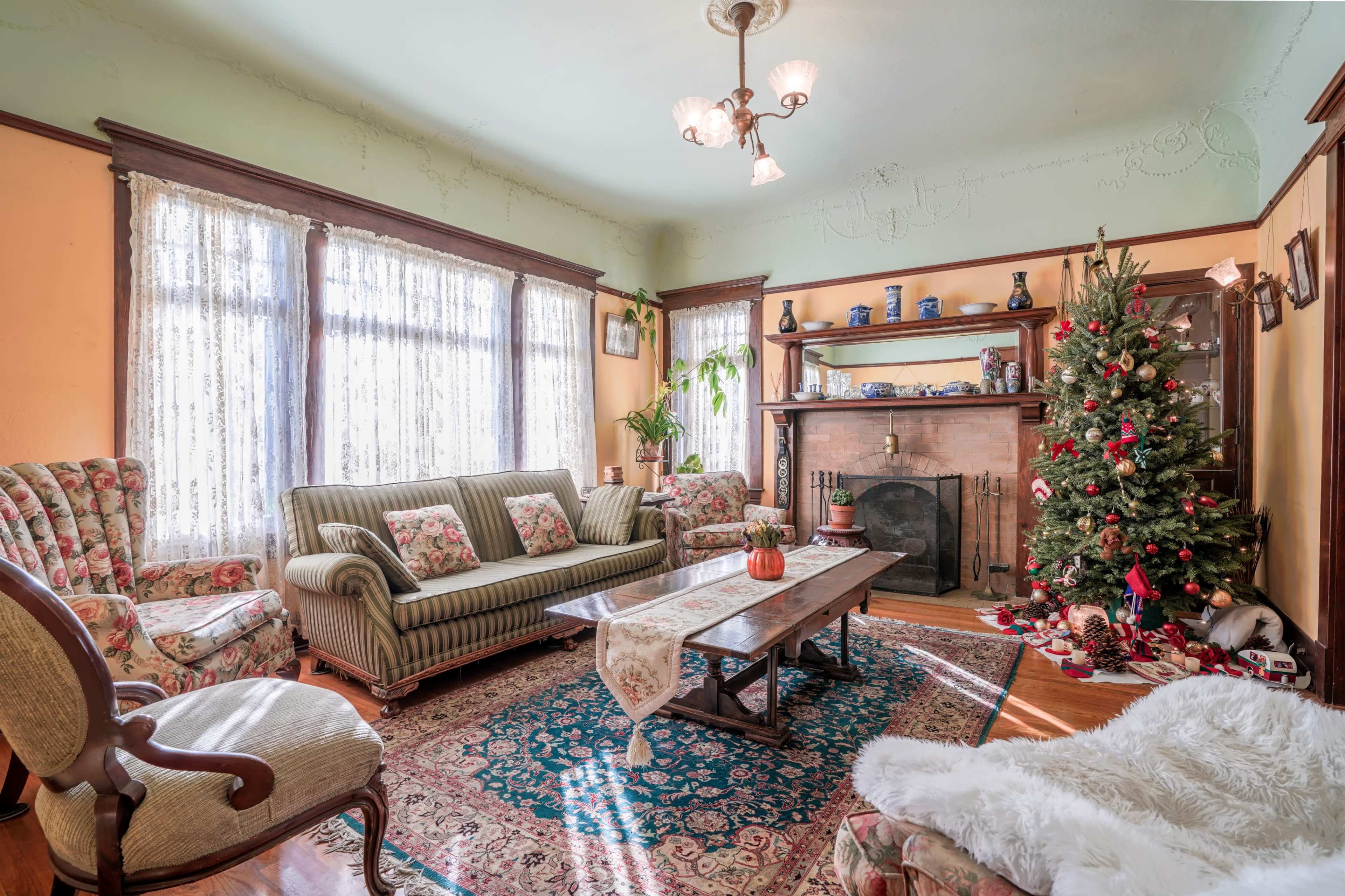 A cozy living room features a patterned sofa, armchairs, a wooden coffee table, and a decorated Christmas tree beside a fireplace, with sunlight streaming through lace curtains.