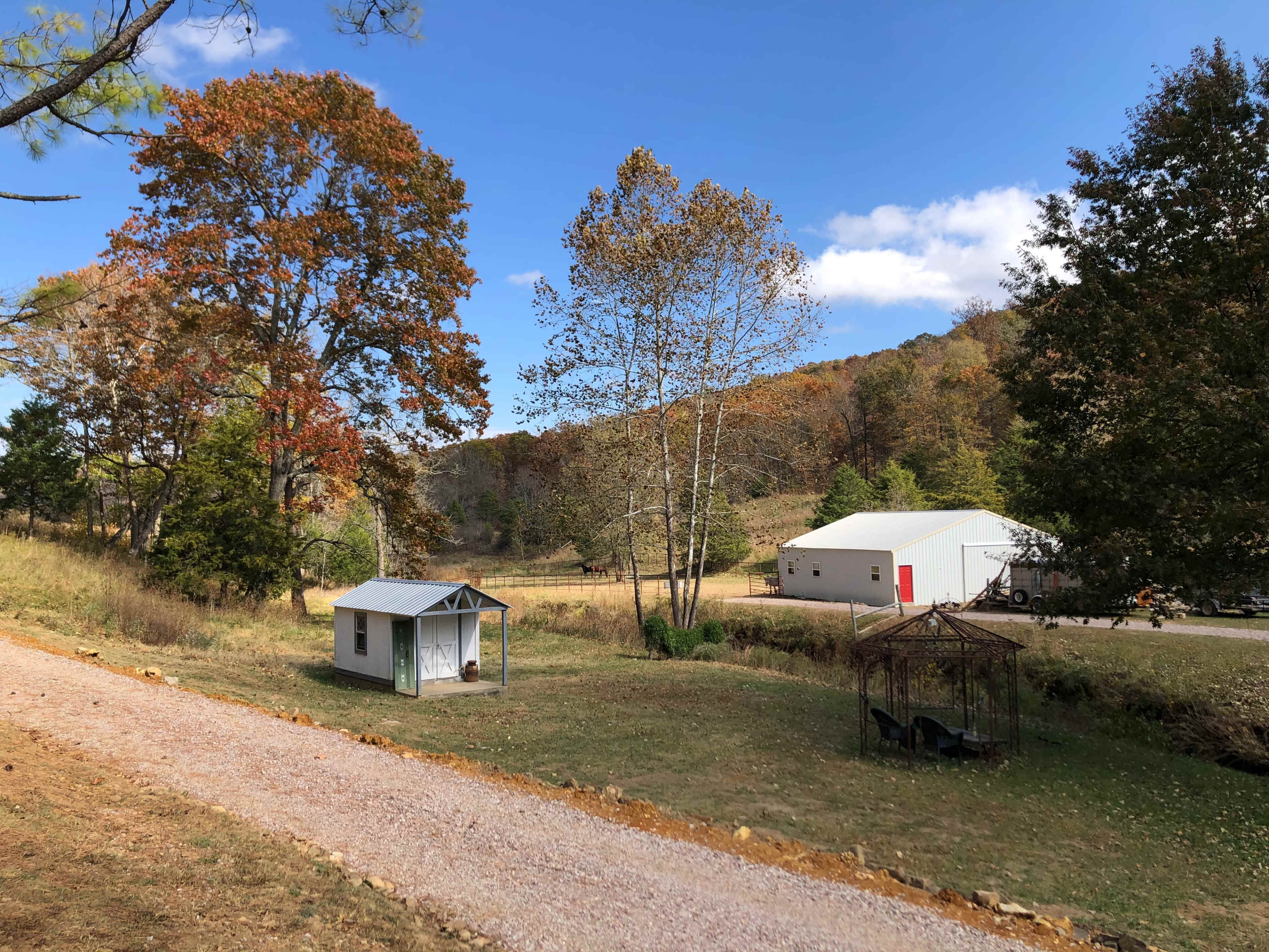 A small shed and a decorative gazebo are set on a grassy area surrounded by trees with autumn-colored leaves and hills in the background.