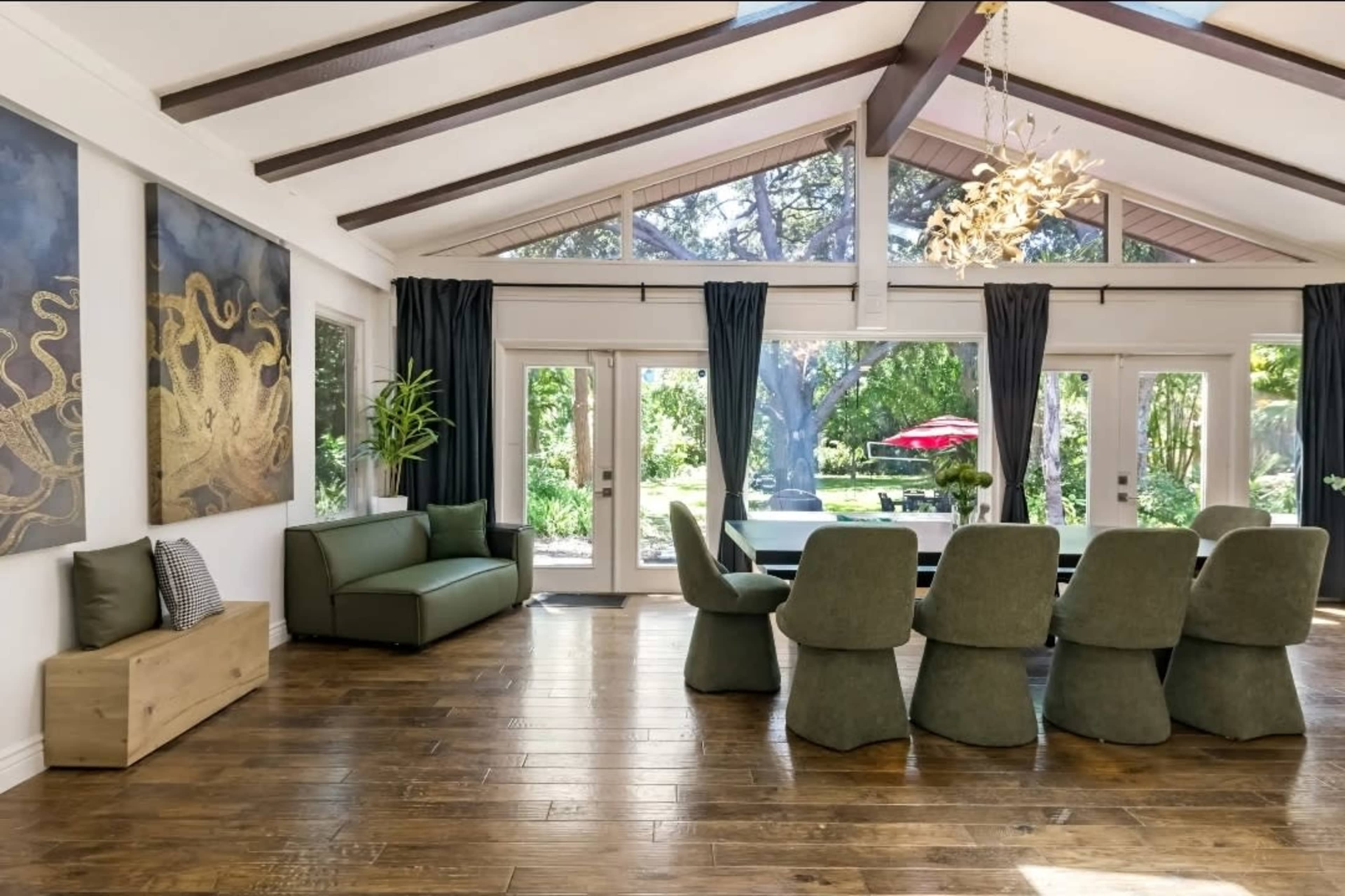The image shows a spacious dining area with a large wooden table surrounded by green chairs, large windows letting in natural light, and a view of a garden outside.