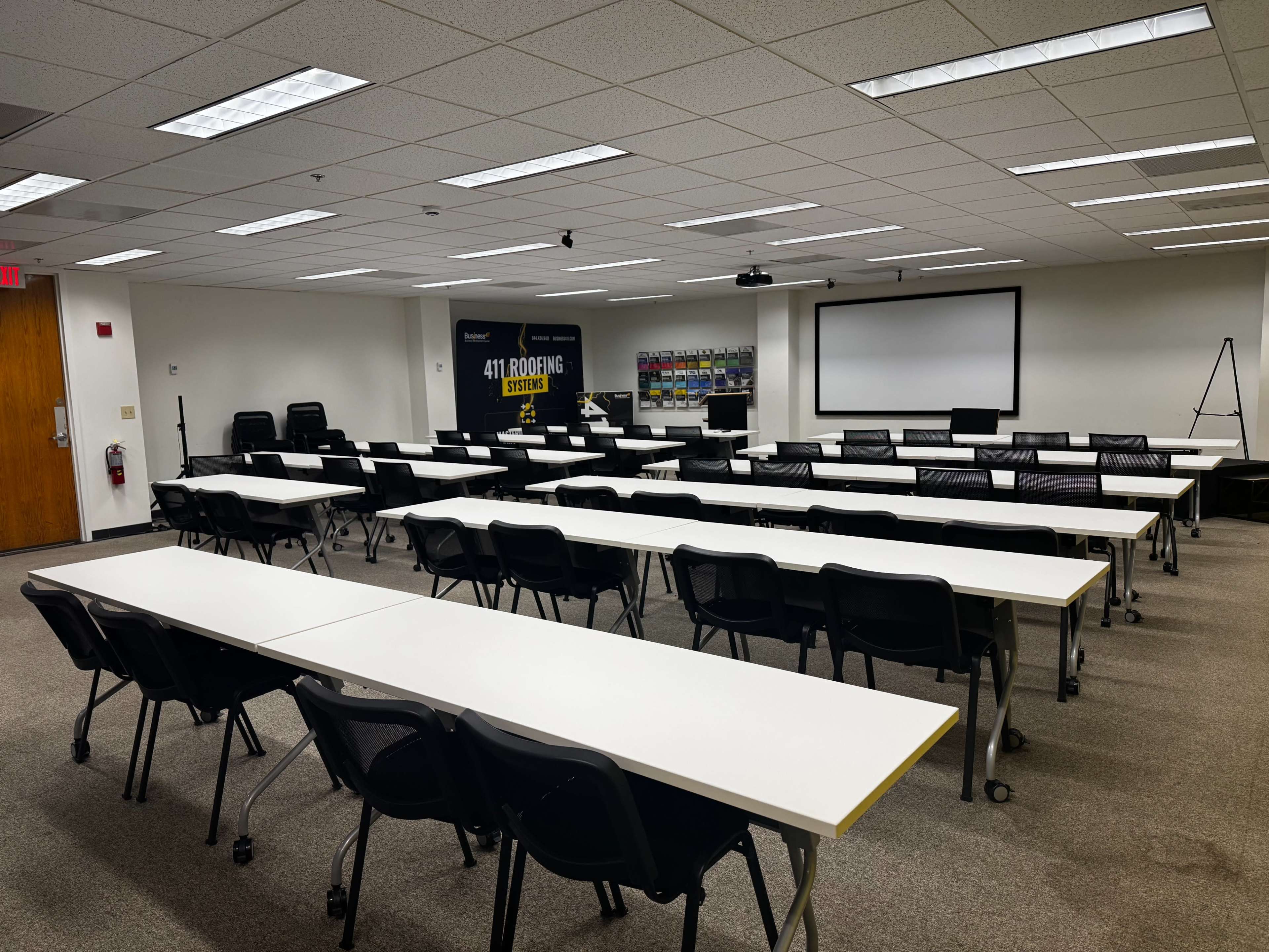 The image shows a meeting room with rows of white tables and black chairs arranged for a presentation, featuring a large screen and a projector at the front.