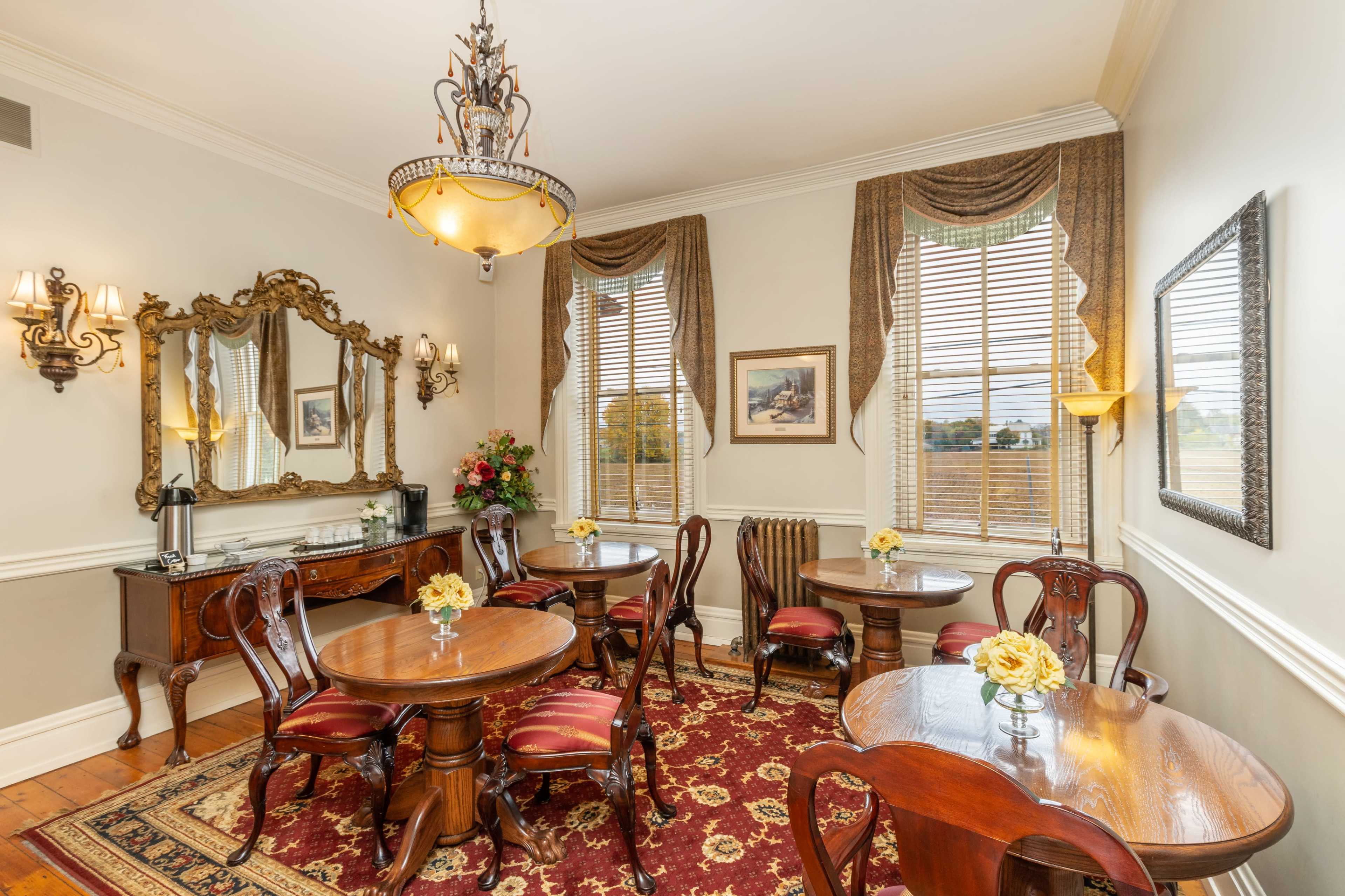 The image shows a cozy dining room with several wooden tables and chairs, adorned with floral centerpieces, a vintage mirror, and large windows dressed with elegant curtains.
